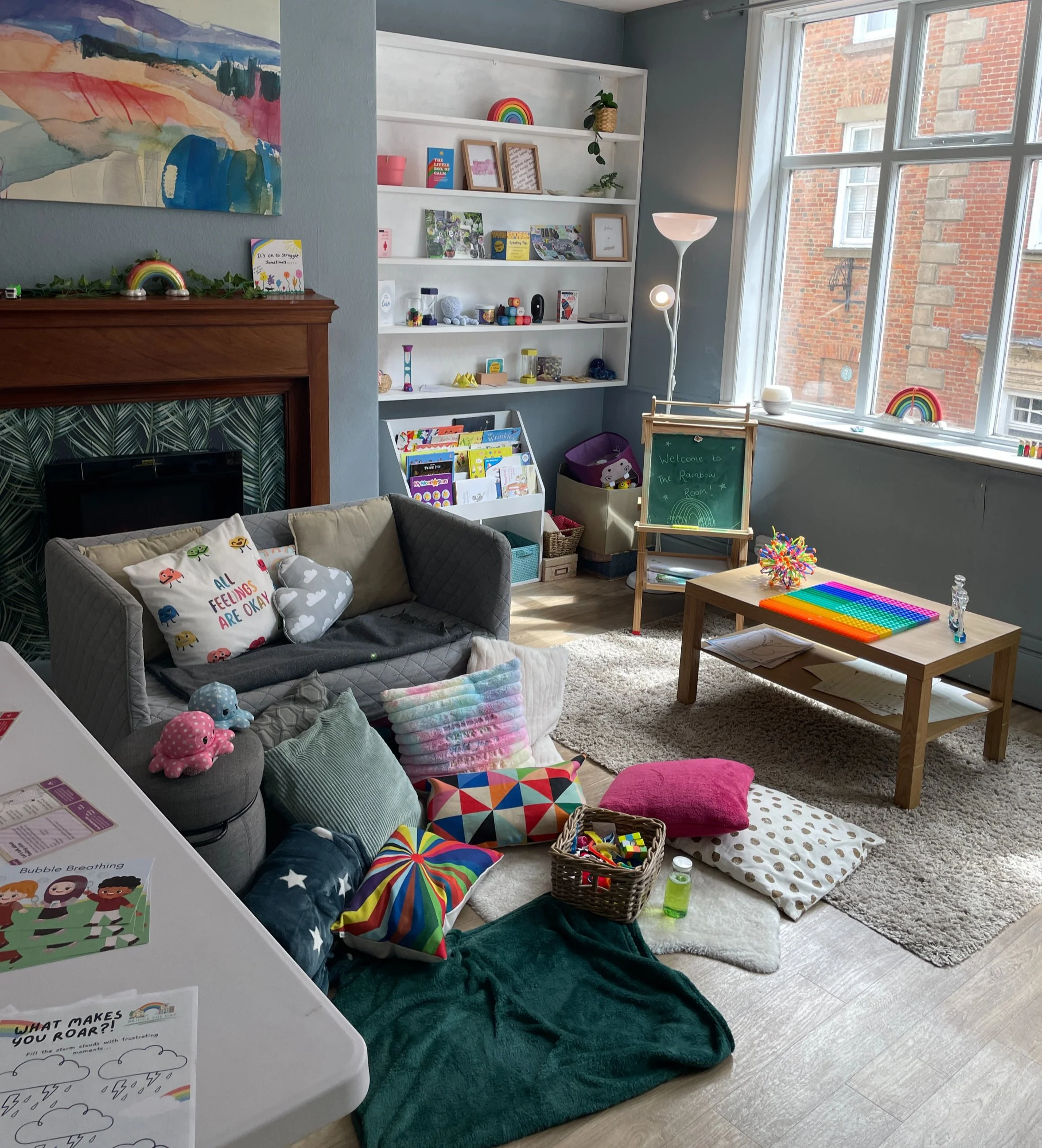 A cozy children's reading and playroom with a gray couch, colorful pillows, a wooden table, a bookshelf filled with books and toys, and a large window letting in natural light.