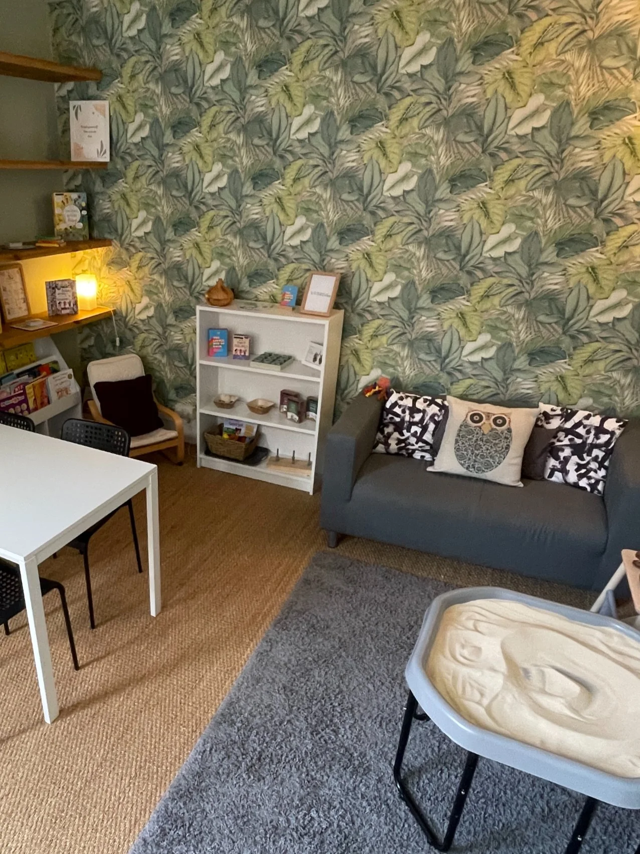 A cozy room with green leafy wallpaper, a gray sofa with owl and patterned cushions, a white bookshelf with books and decorative items, a white table with black chairs, a corner with a small armchair and shelves, and a table with a baby changing pad 