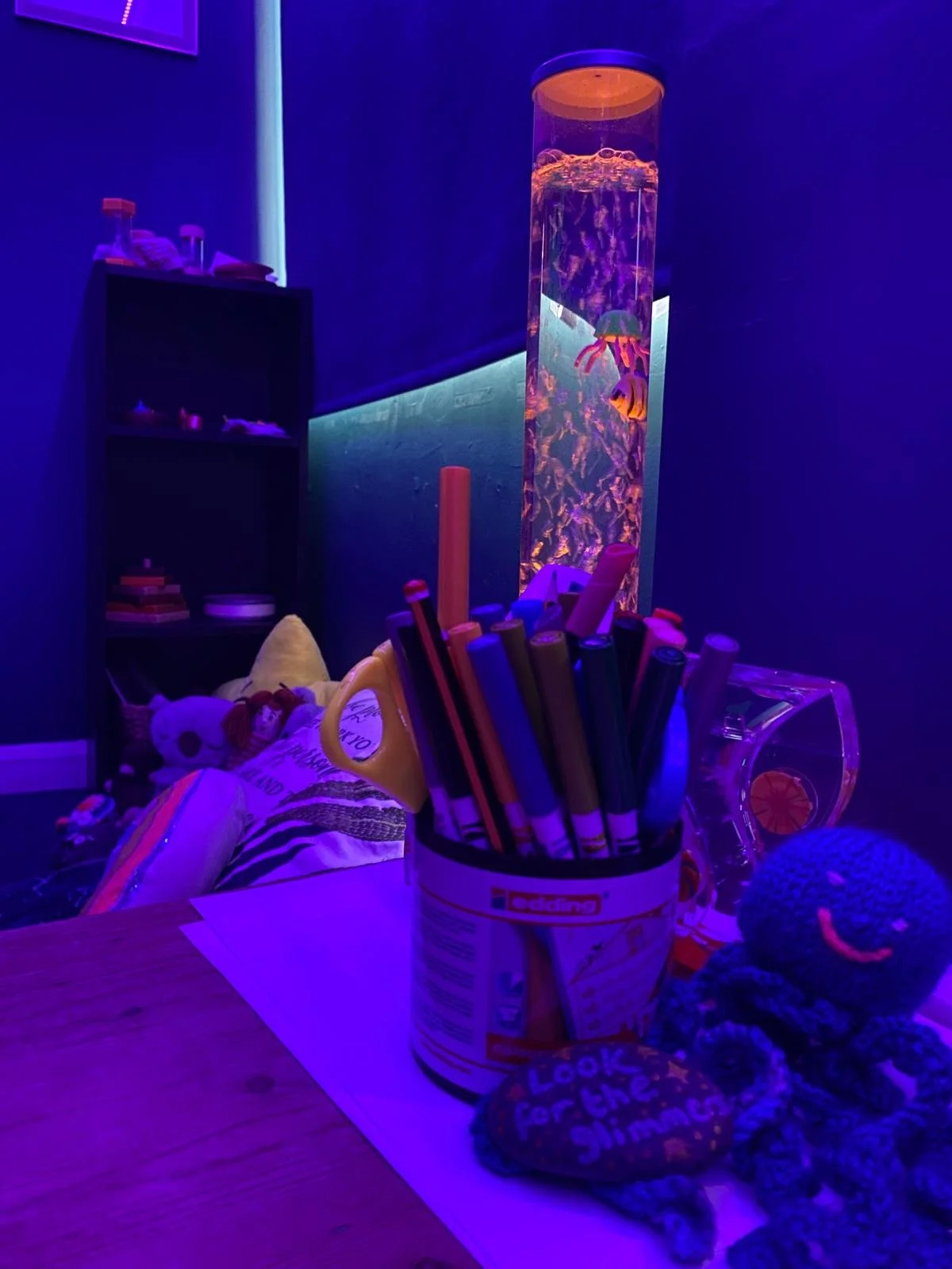 A dark room illuminated with purple and blue lights, containing a tall cylindrical fish tank with fish, a black shelf with small objects, a stuffed doll, a container with colored markers, and a crocheted octopus toy on a wooden table.