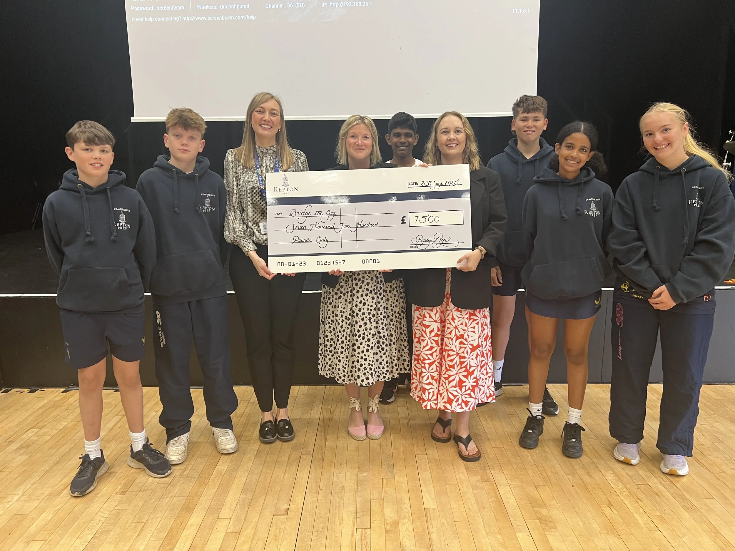 A group of nine children and three adults holding a large check on a stage. The check is made out for 7,500 pounds and is signed by Repton. The group is smiling and dressed casually, some wearing hoodies with a logo. The background features a blank p