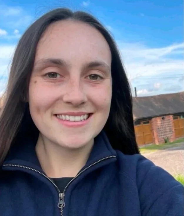A young woman with long dark hair smiling outdoors, wearing a navy zip-up jacket, with a wooden fence and a building in the background, under a blue sky with some clouds.