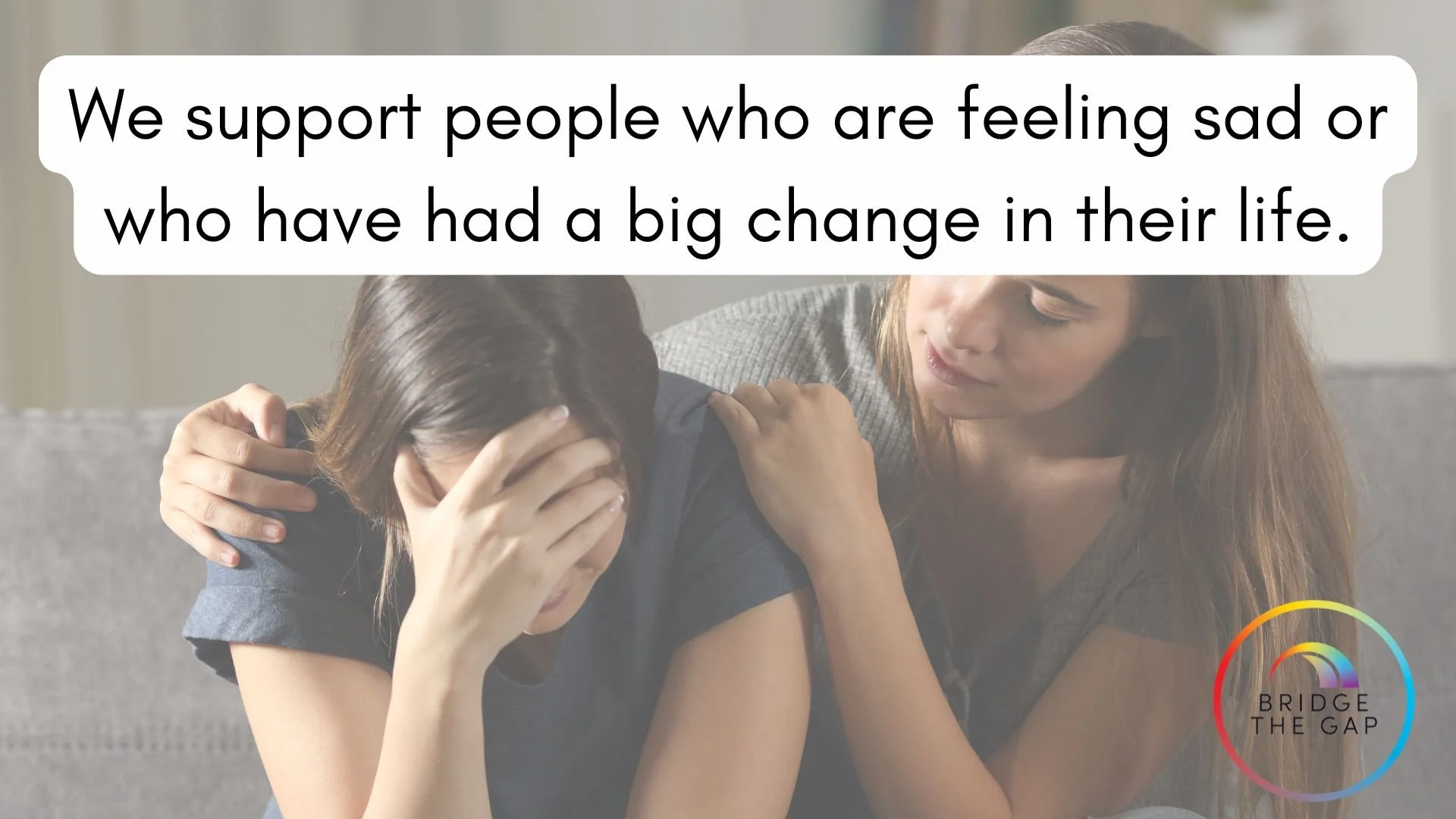 A woman comforting another woman who appears distressed, with a supportive gesture, sitting on a sofa.