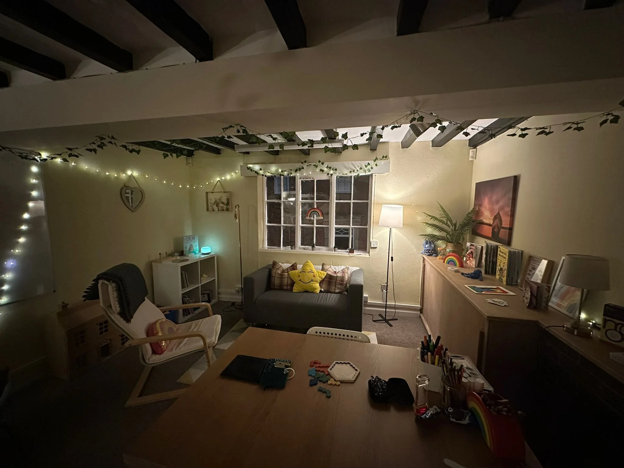 Living room with string lights and garland hanging from the ceiling, a window with a rainbow decoration, a lamp, a bookshelf with books and decor, a gray couch with plaid pillows and a yellow smiley face pillow, a wooden sideboard with books, plants,