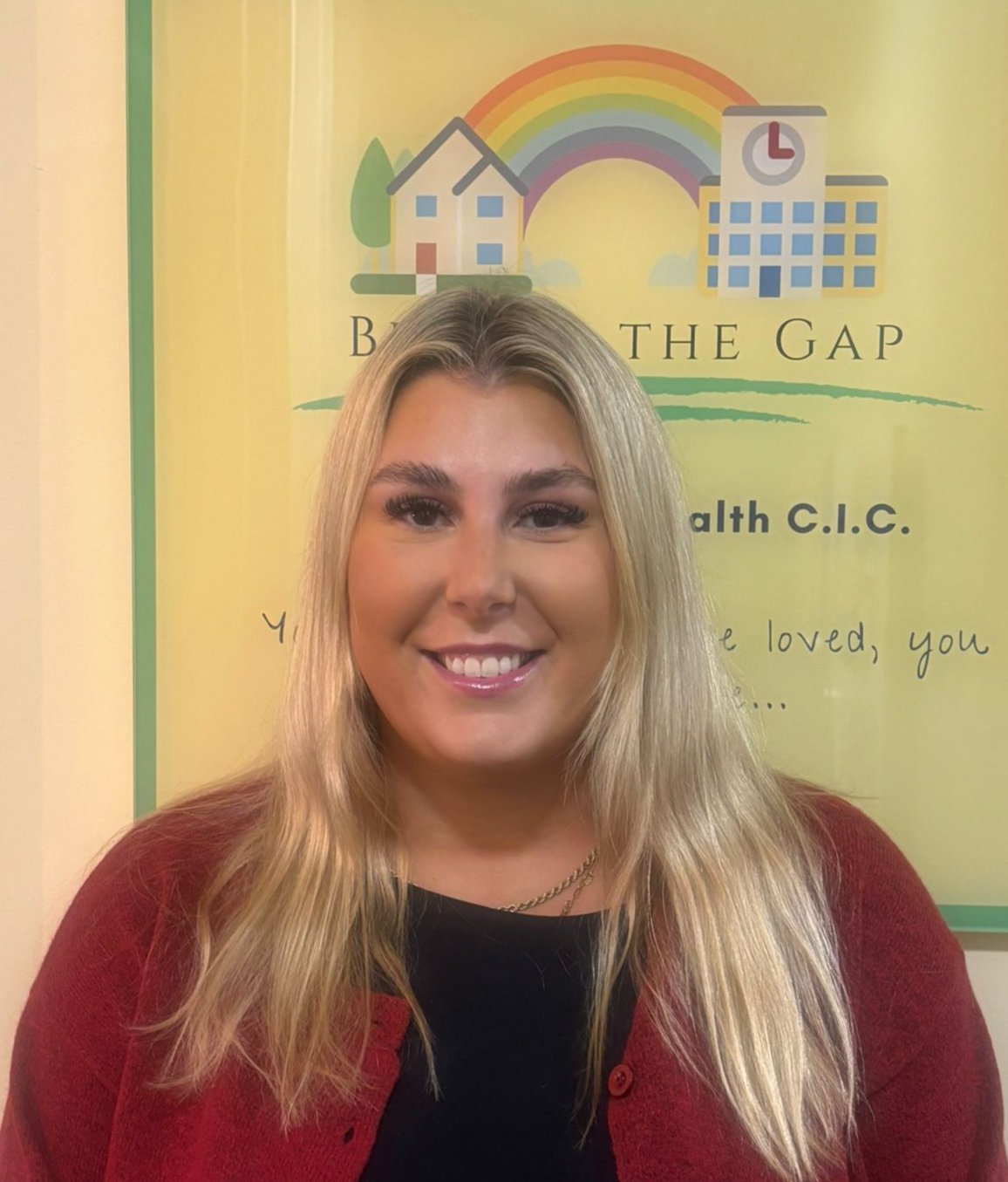 Smiling young woman with long blonde hair, wearing a black top and red cardigan, standing in front of a yellow poster with rainbow, house, and clock images, and the text 'BREAK THE GAP' and 'Health C.I.C.'
