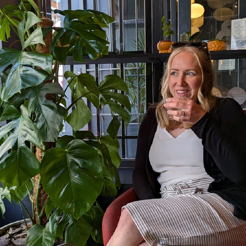 A woman sitting indoors next to large green plants, holding a small cup near her mouth, with a window and decorative items in the background.