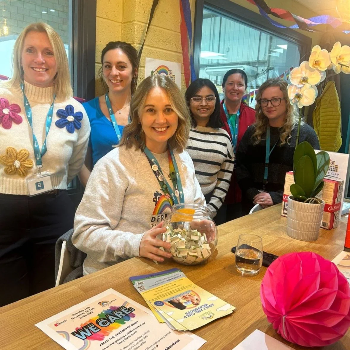 Six women gathered around a table decorated with colorful rainbow-themed decorations, including paper flowers and banners. One woman seated at the table is holding a glass fishbowl filled with small paper pieces, and there are pamphlets and a potted 