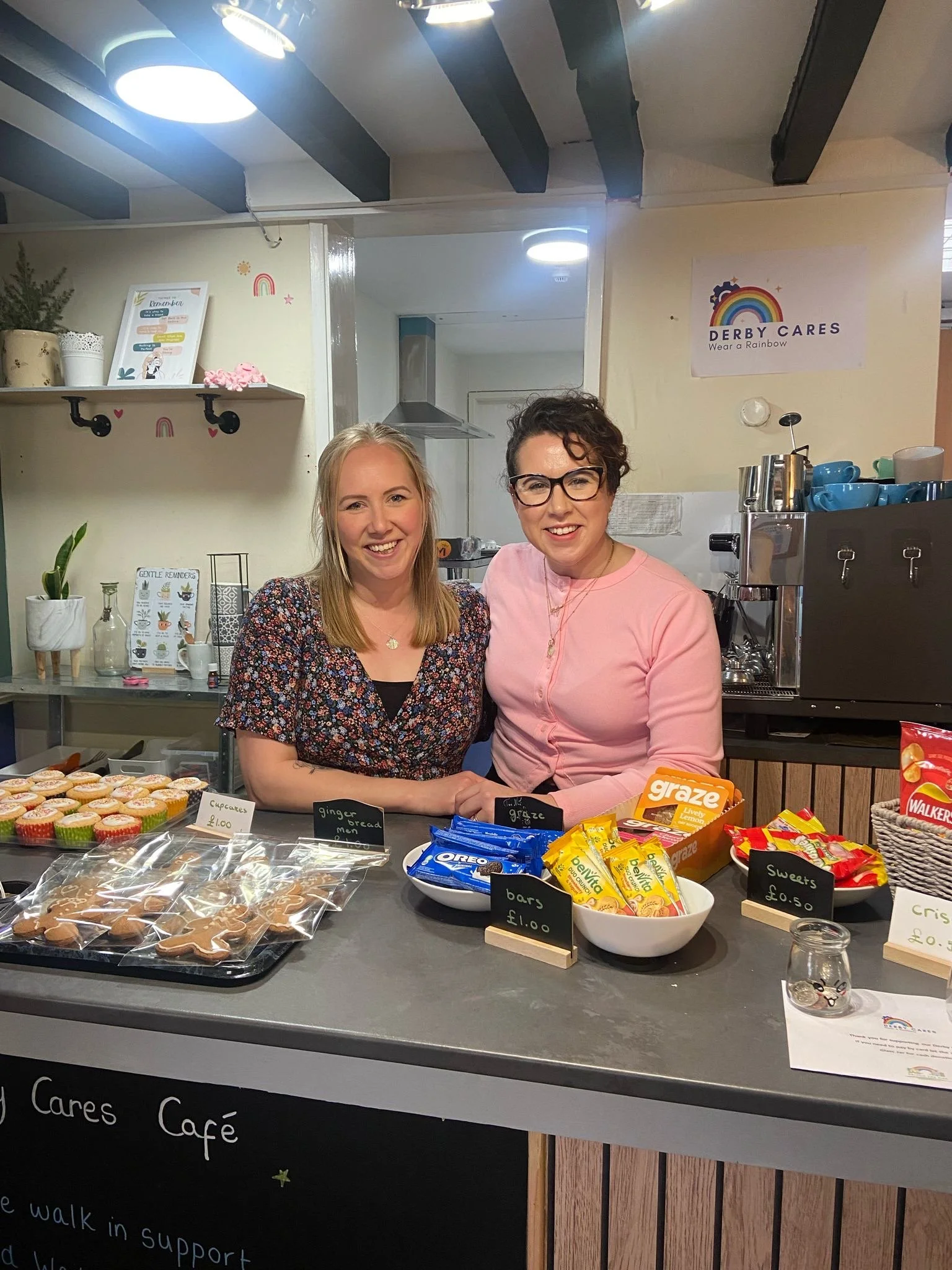 Two women standing behind a counter at a café, smiling, with various baked goods and candy on display.