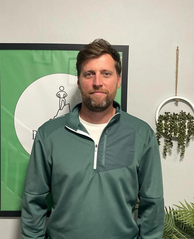 A man with a beard and short brown hair smiling, wearing a gray button-up shirt and a colorful lanyard, sitting indoors against a green wall with framed pictures.