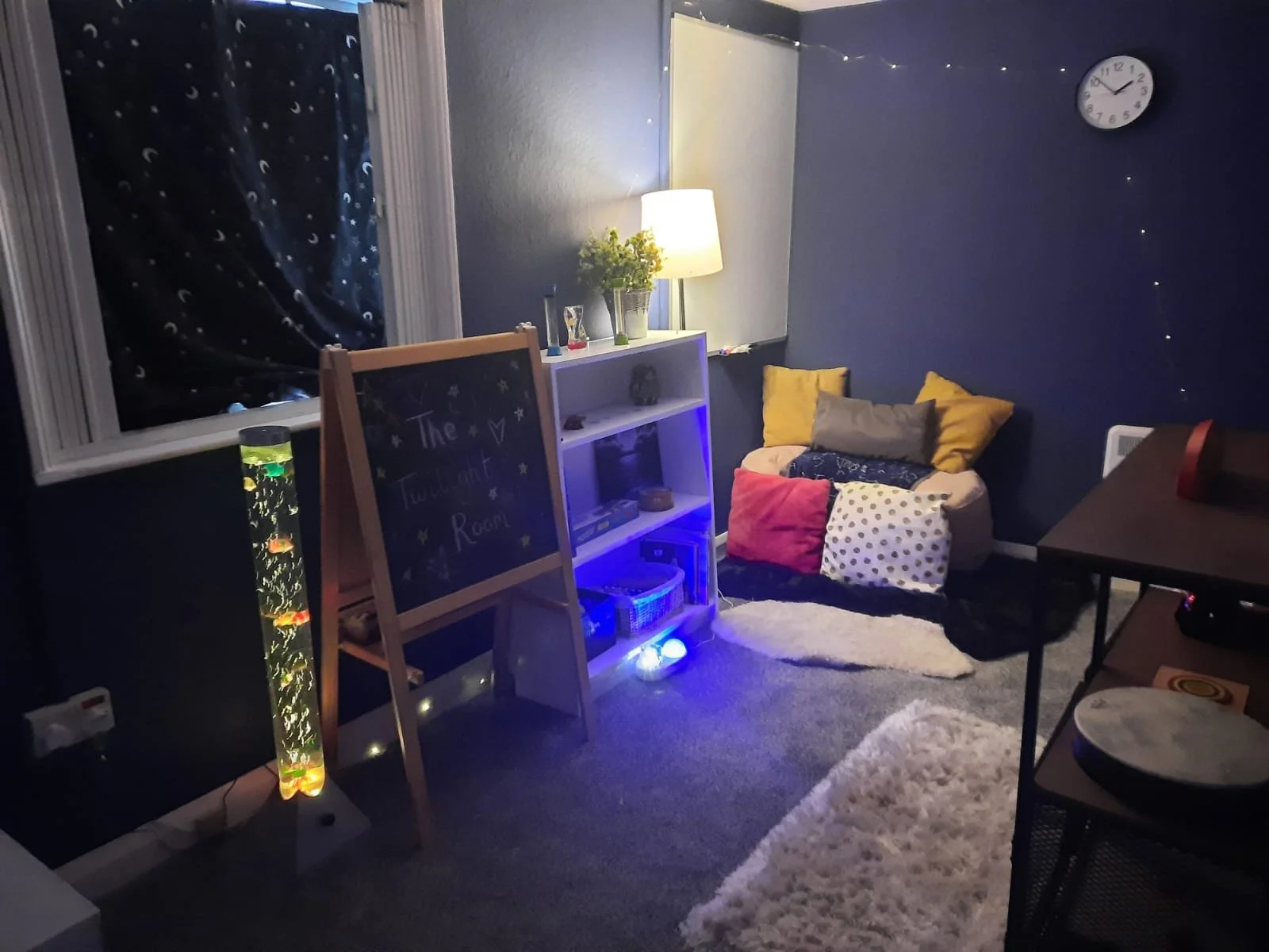 Cozy room with a white bookshelf, pillows on a bean bag, a lamp, a clock on the wall, a lava lamp, and fairy lights against dark blue walls.