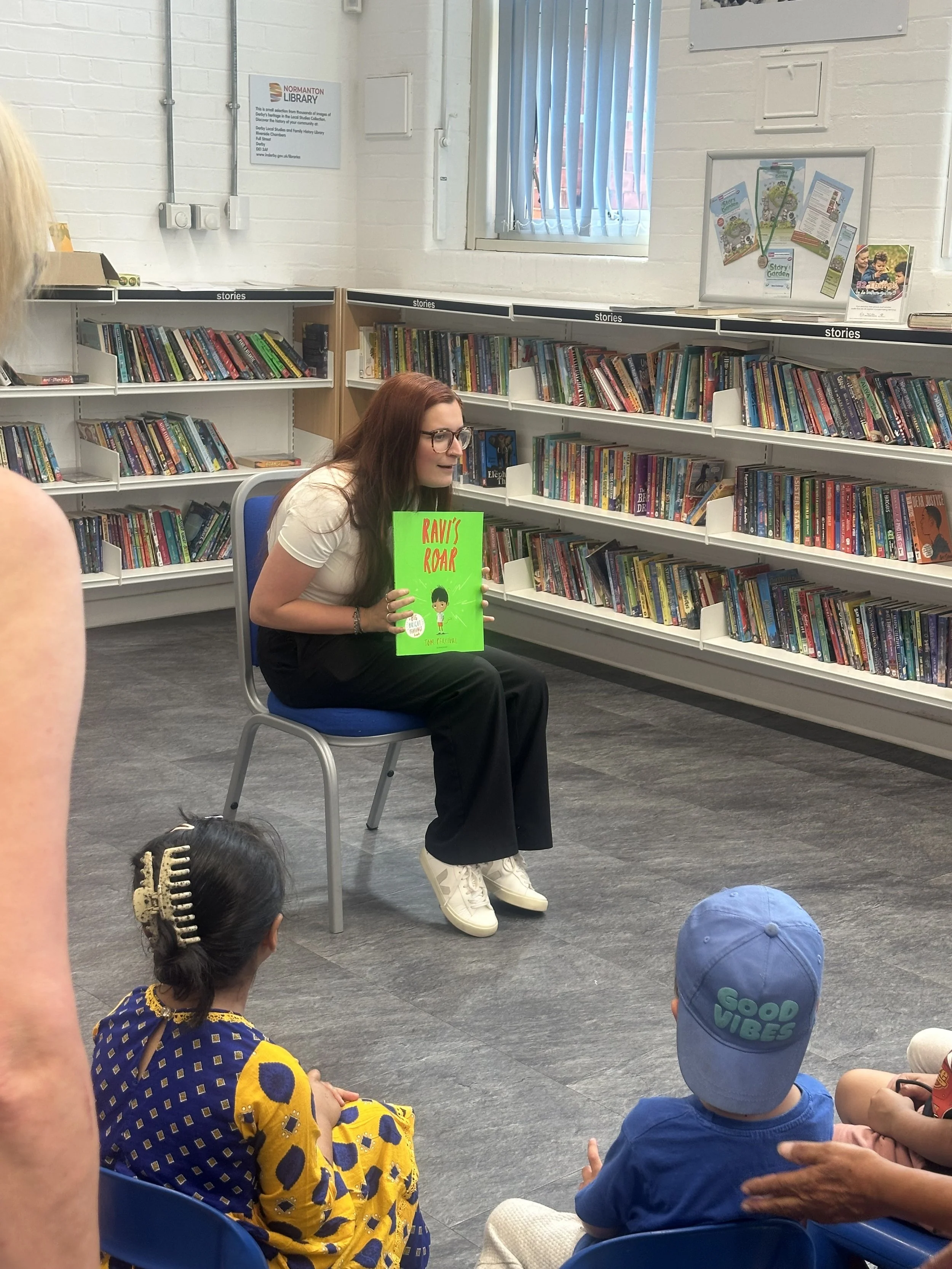A woman with glasses and long hair is sitting on a blue chair in a library, reading a children's book titled "Ravi's Roar" to children seated on the floor. The library shelves are filled with colorful books.