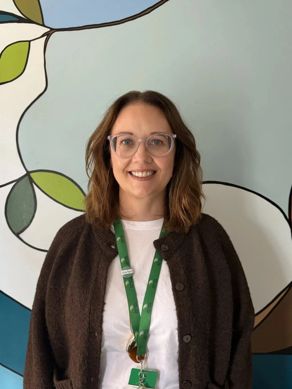 A woman with shoulder-length blonde hair, wearing a black blazer over a white top, sitting on a beige couch with green and white patterned pillows, smiling at the camera in a cozy indoor setting with wooden and frosted glass wall panels in the background.