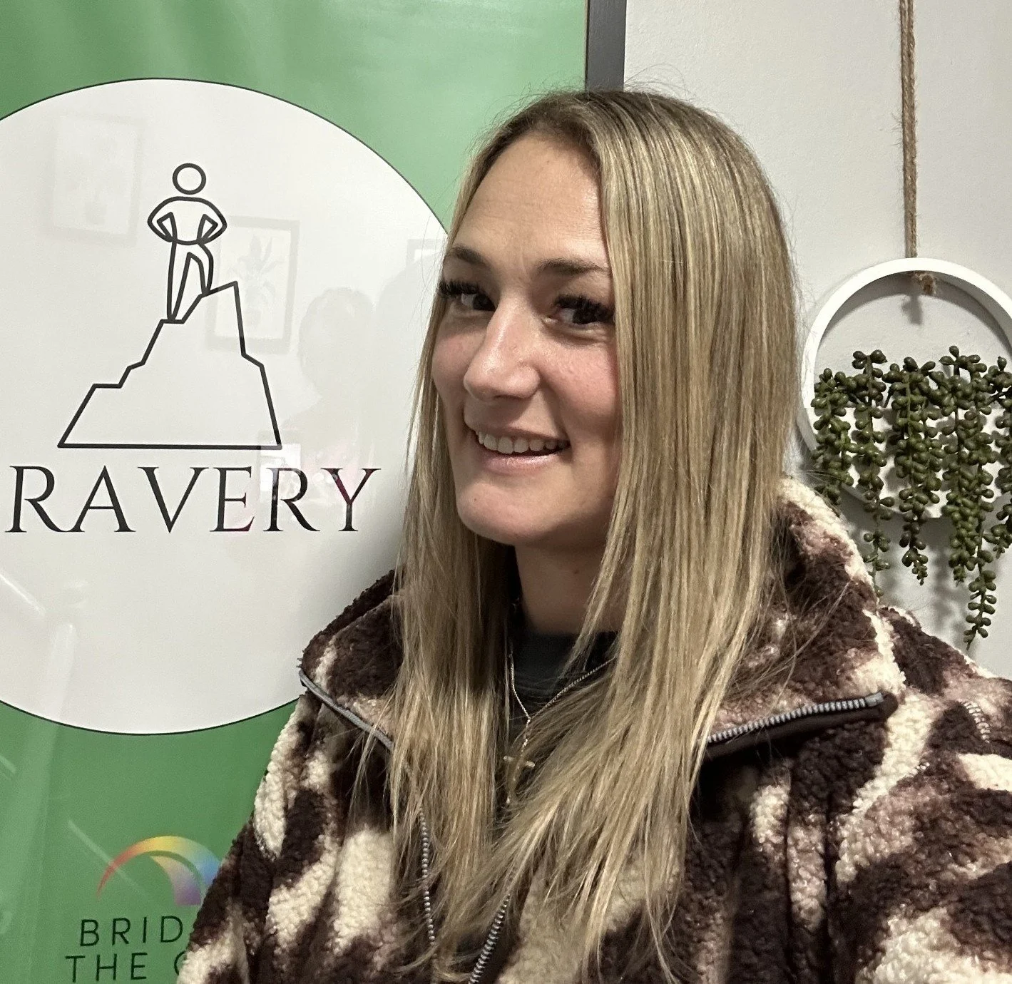 A young woman with long blonde hair smiling at the camera, wearing a brown and cream patterned fleece jacket, standing in front of a sign that says 'RAVERY' with a logo of a person standing on a mountain.