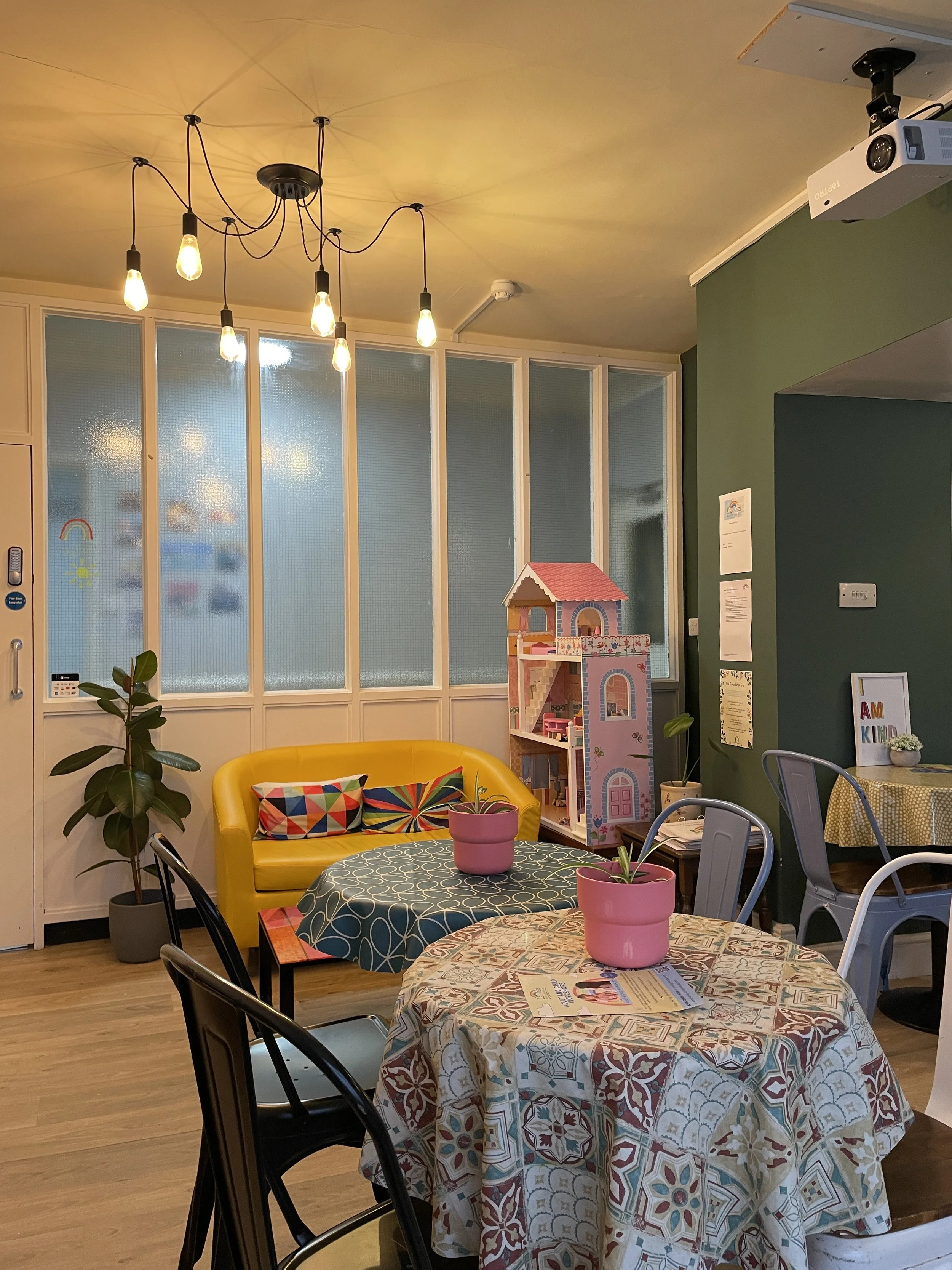 Cozy indoor space with a yellow couch, colorful pillows, a round table covered with a patterned tablecloth, and pink potted plants. Background includes a dollhouse, large potted plant, and textured glass partitions, with warm lighting from hanging bu