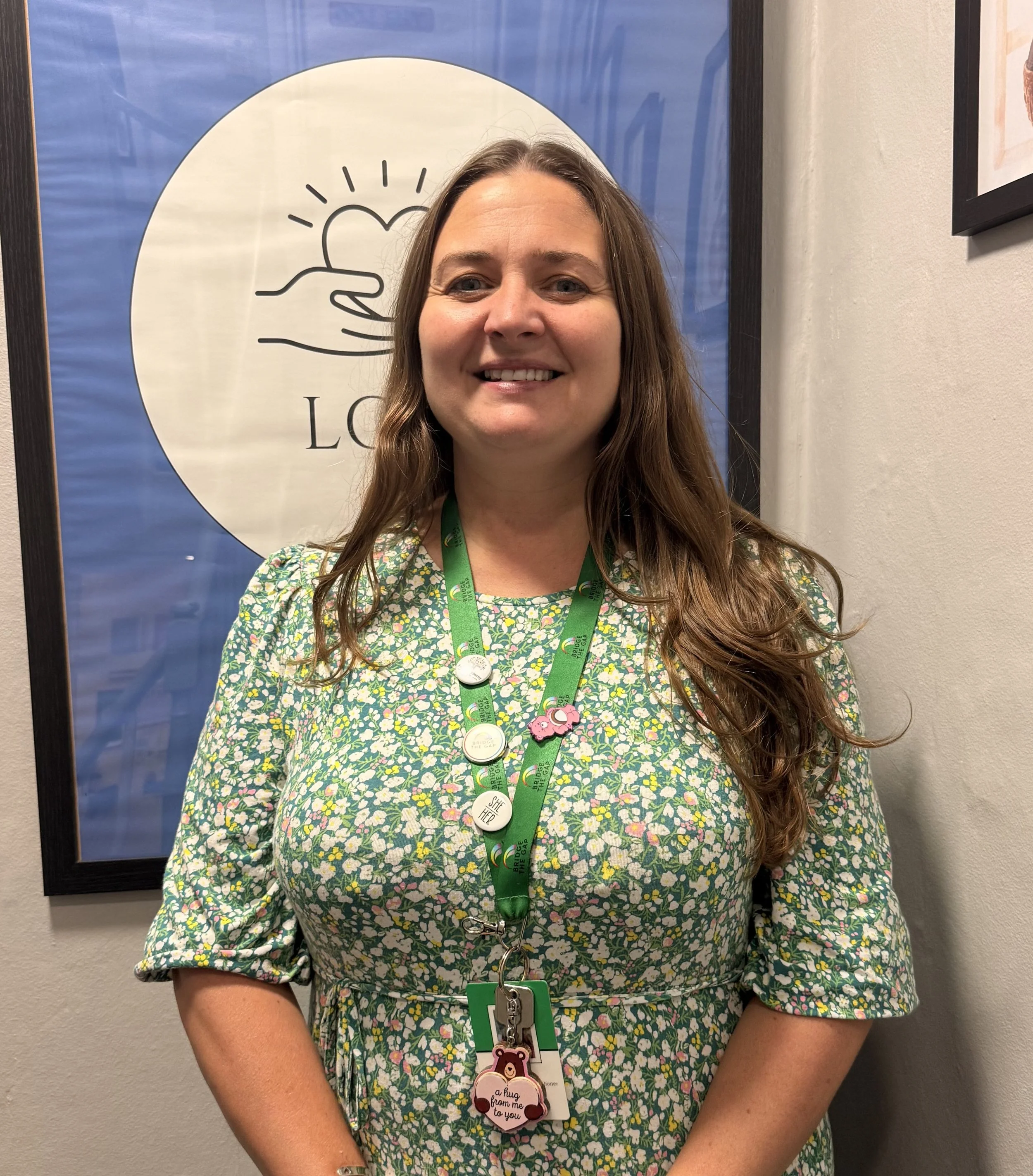 A woman with long brown hair, wearing a pink top, smiling, with a colorful lanyard with pins around her neck, standing in front of a leafy green and pink floral patterned wall.