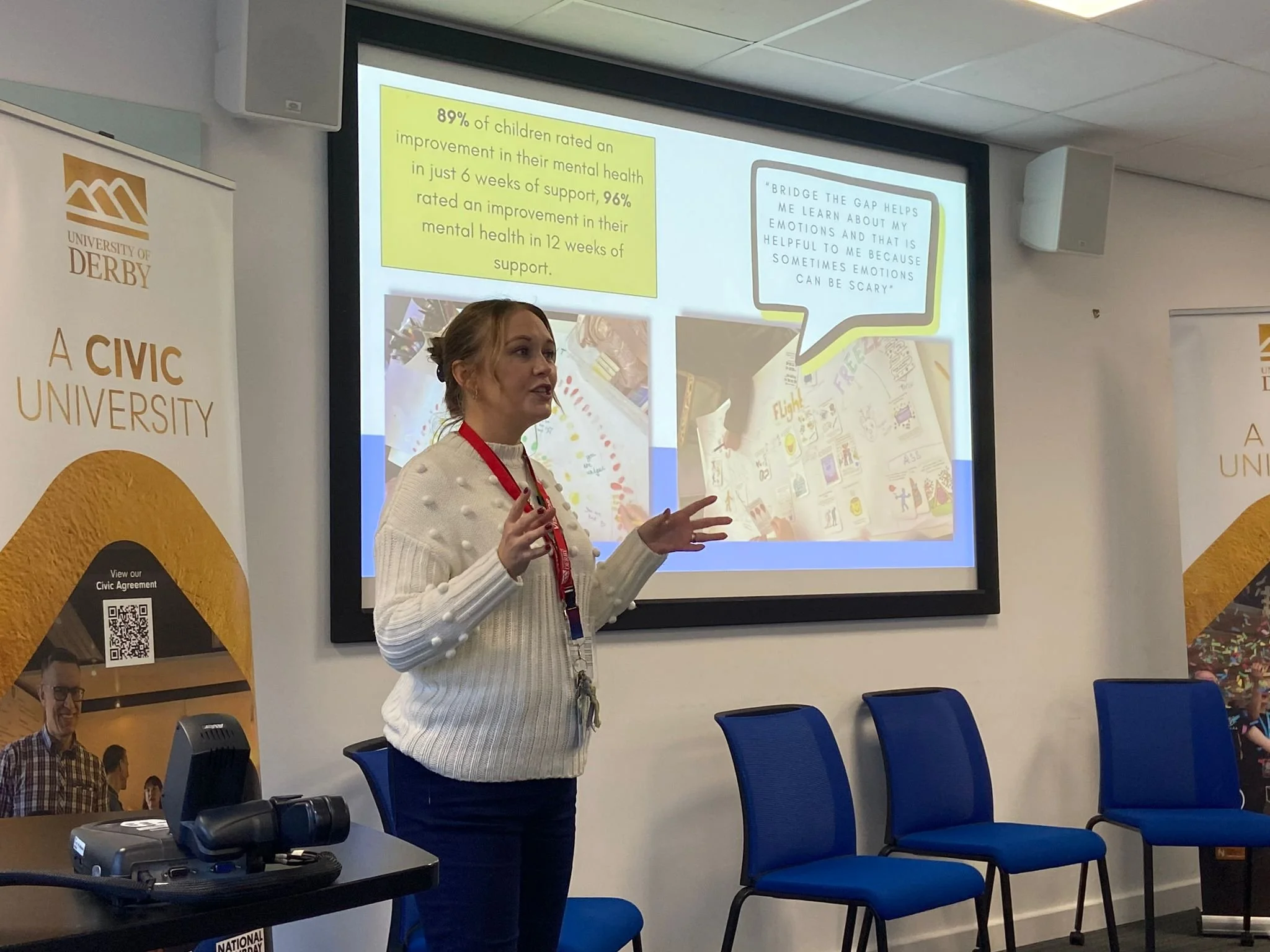 A woman presenting in front of a screen with a slide, at University of Derby, with blue chairs arranged along the wall.
