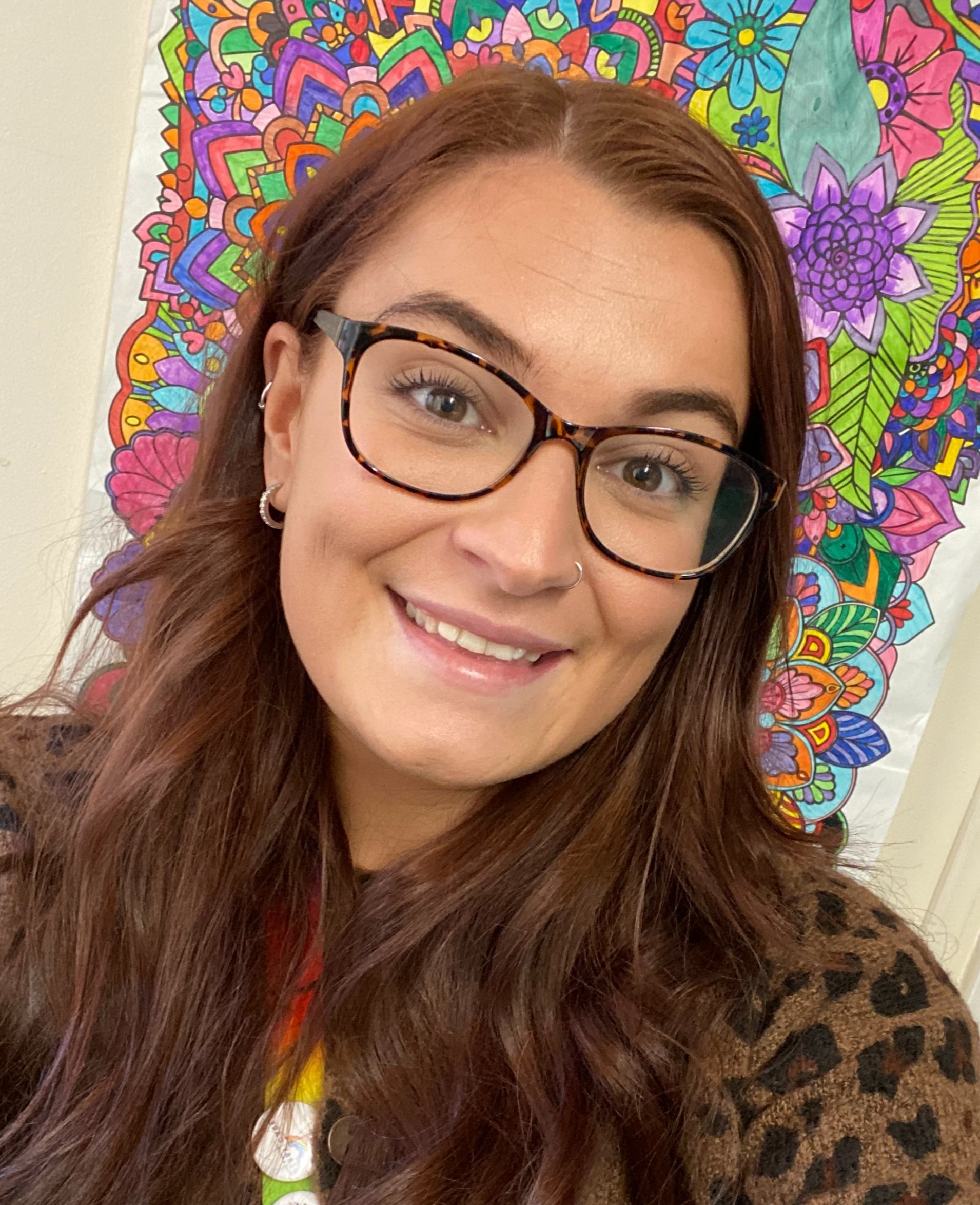 A young woman with long, wavy brown hair, wearing glasses with tortoiseshell frames, smiling, and standing in front of a colorful, intricate mandala design on a wall.