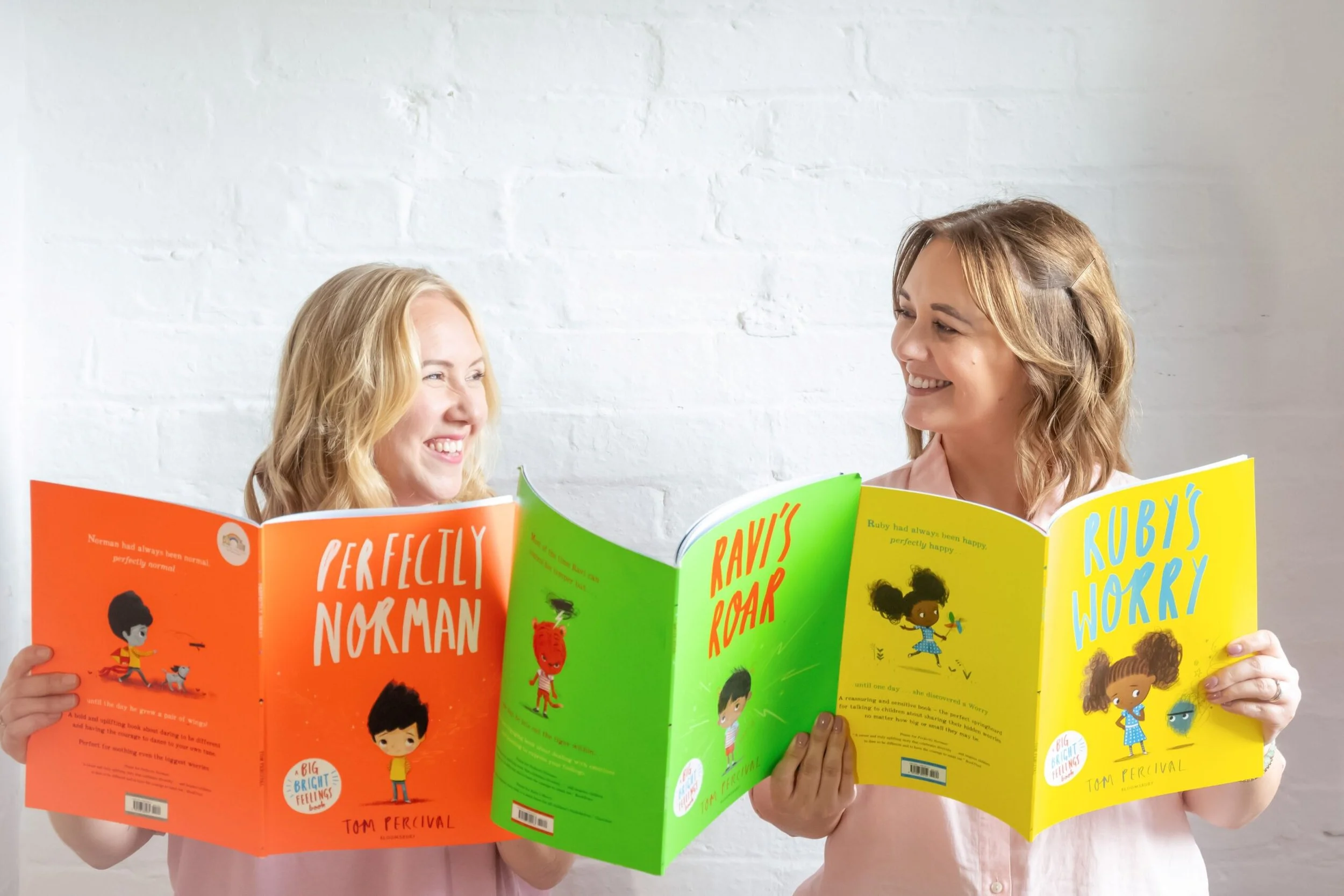 Two women smiling and holding colorful children's books in front of a white brick wall.