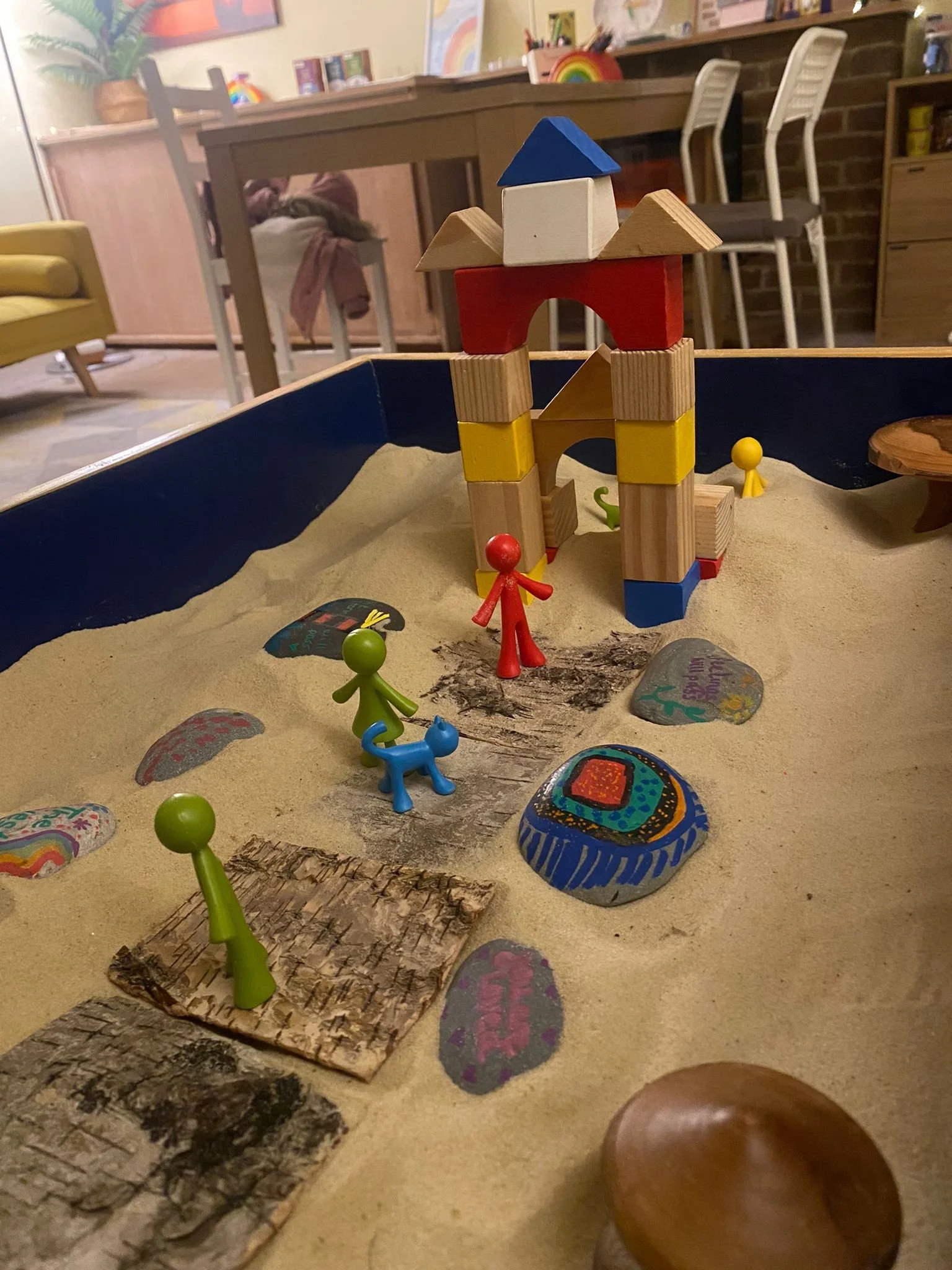 Colorful toy castle with toy figures and painted rocks on a sandy surface, with furniture and decorations in the background.