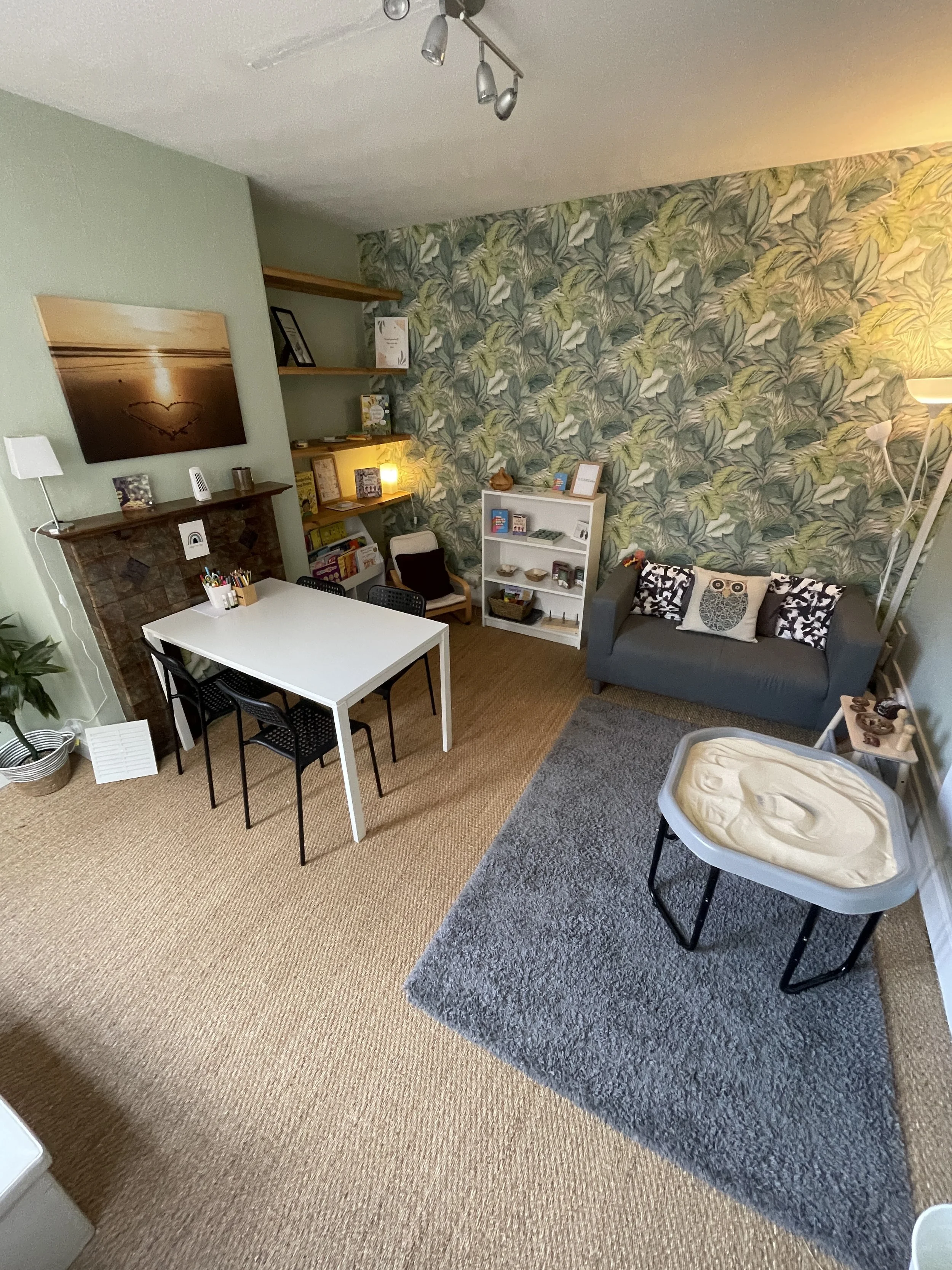 A cozy living room with a green wall, a gray sofa with decorative pillows, a white bookshelf, a dining table with four chairs, a large sunset photograph, a floor lamp, and woven carpet flooring.