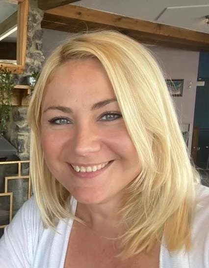 A woman with shoulder-length blond hair smiling in a cozy indoor setting.