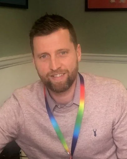 A man with a beard and short brown hair smiling, wearing a gray button-up shirt and a colorful lanyard, sitting indoors against a green wall with framed pictures.
