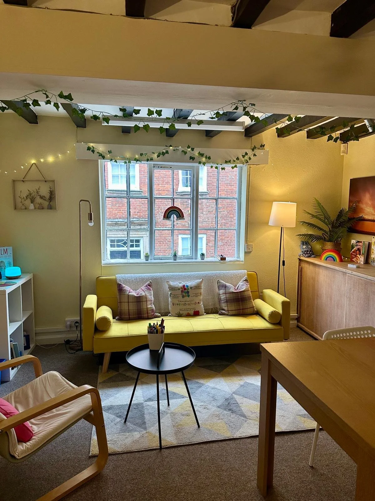 A cozy living room with a yellow sofa, plaid cushions, and a small black round table. There is a large window with small potted plants and a rainbow decoration hanging inside. The room has green ivy string lights and a floor lamp, with a sideboard de