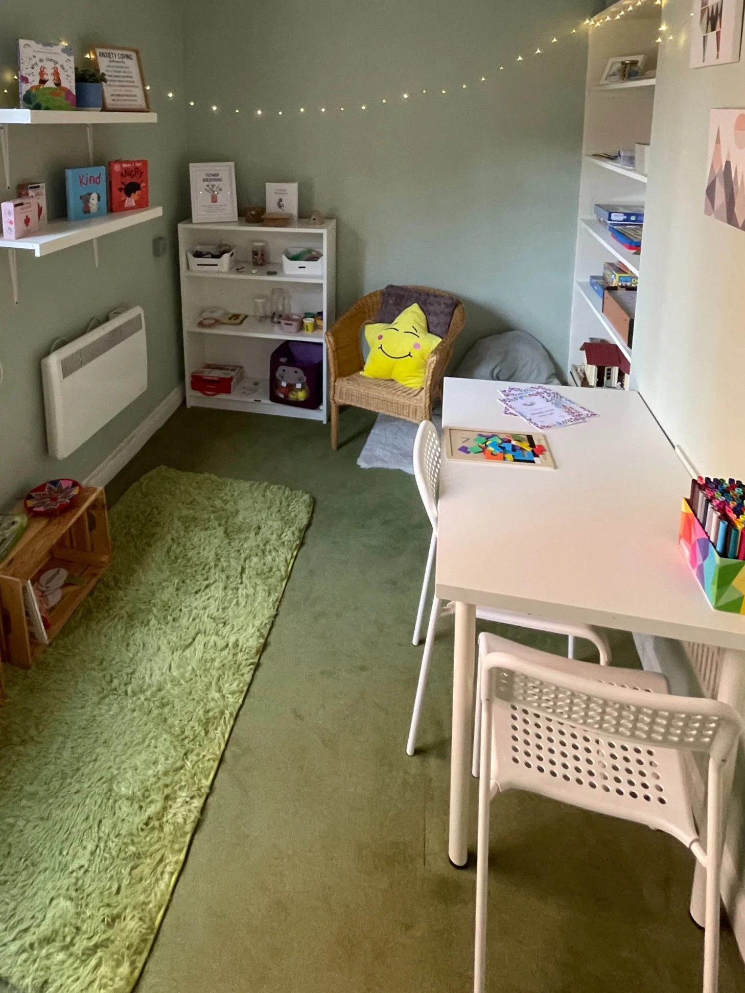 A cozy children's playroom with a white table and chairs, a green shag rug, a wicker chair with a yellow smiley face pillow, white shelves filled with toys and books, a string of fairy lights on the wall, and framed artwork and posters.