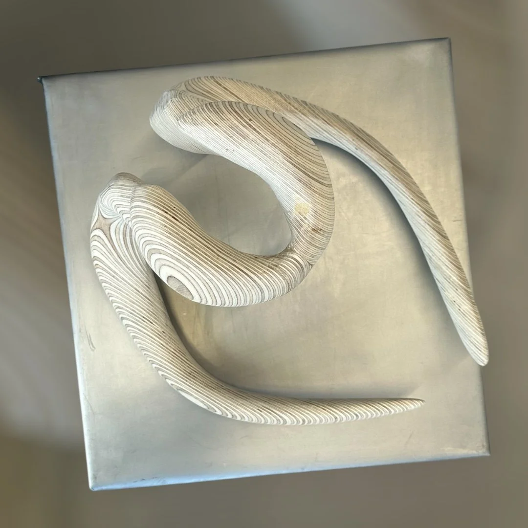 A wooden, abstract sculpture with curved, layered patterns on a metallic square base.