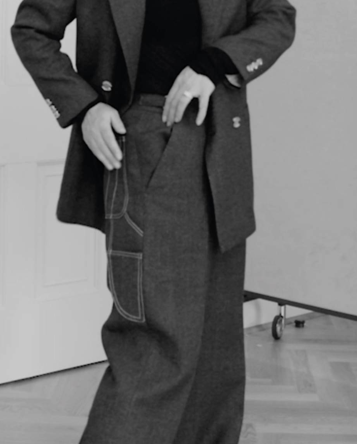 Black and white photo of a person dressed in formal attire, standing indoors with their hands on their hips.