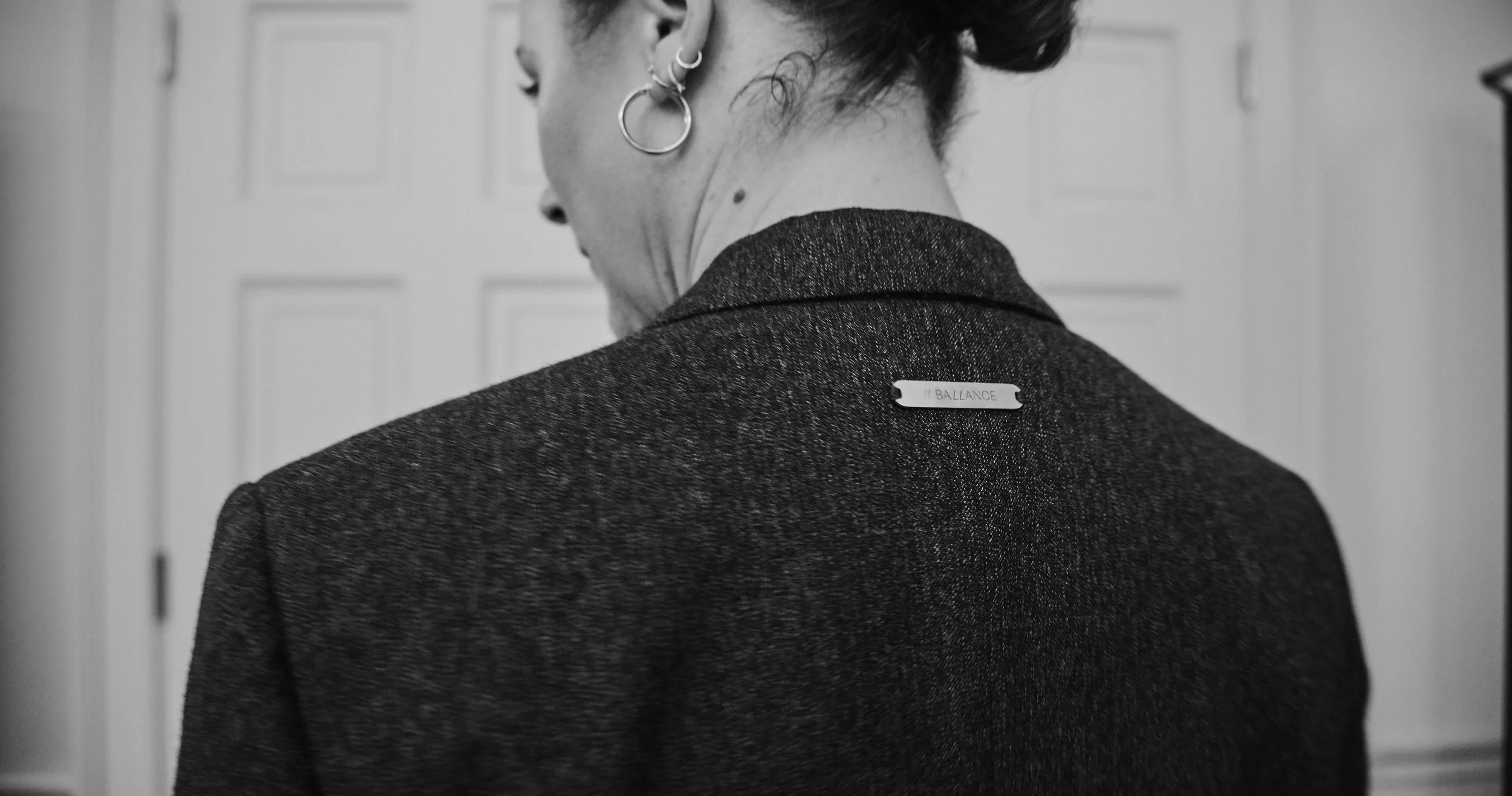 A black-and-white photo of a woman from behind, wearing earrings and a dark blazer with a small label reading 'BALANCE' on the shoulder.