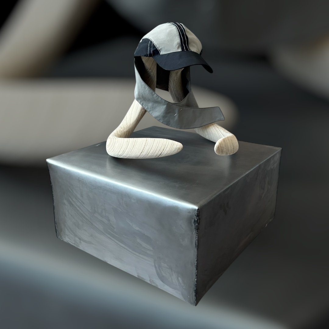 A wooden sculpture resembling a person with arms and legs, wearing a black and white cap and a black and gray visor, displayed on a gray pedestal in a gallery.