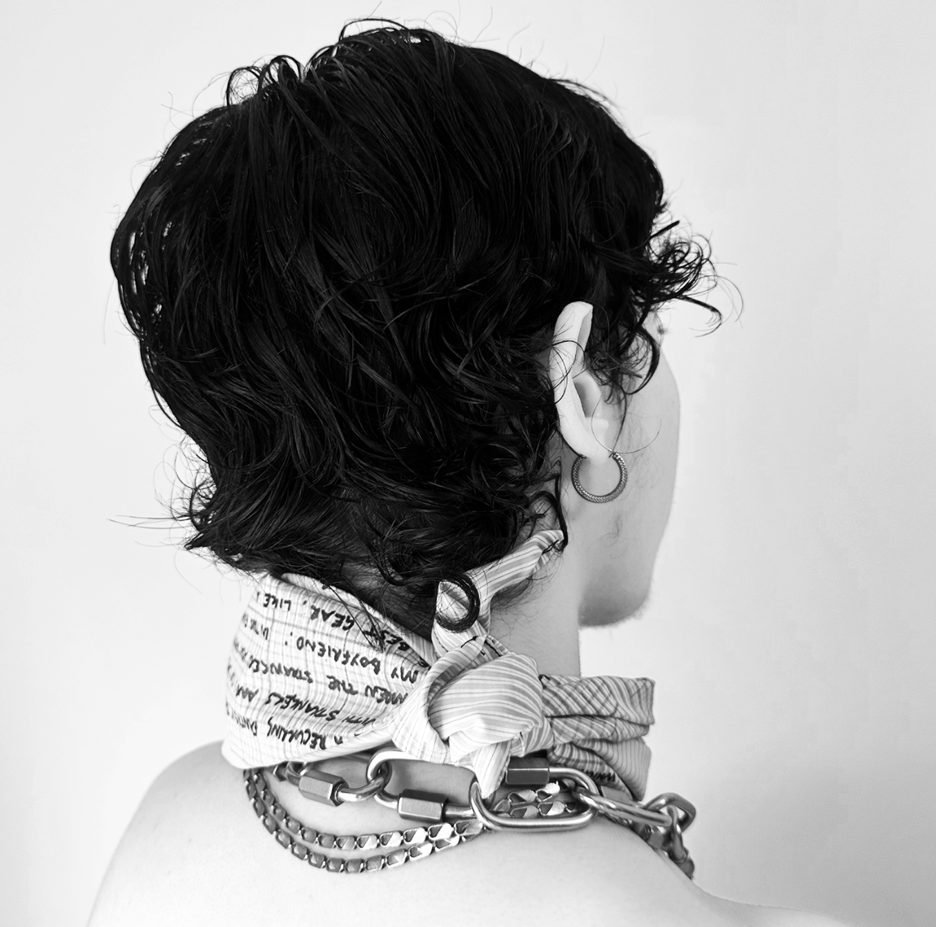 Back of person with short curly dark hair, wearing a chain necklace, hoop earring, and a patterned scarf tied around the neck, against a plain background.