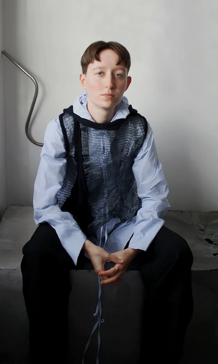 A young person with short brown hair, wearing a white button-up shirt and a black mesh vest, sits on a metal surface against a plain white wall, looking at the camera.