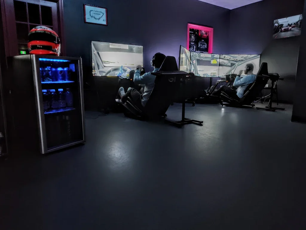 Two individuals are sitting in racing simulator chairs with multiple monitors displaying a racing game or simulation. A beverage fridge with blue lighting is on the left, and a racing helmet is on top of it. The room has dark walls, with framed pictures and a window or painting, creating an immersive racing or gaming experience.