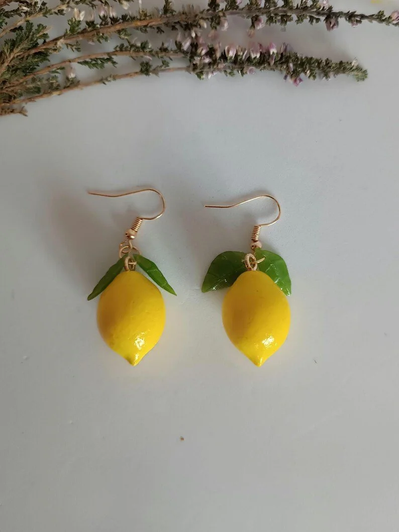 Handmade yellow polymer clay lemon earrings with realistic green leaf details. Fresh summer fruit jewelry flat lay.