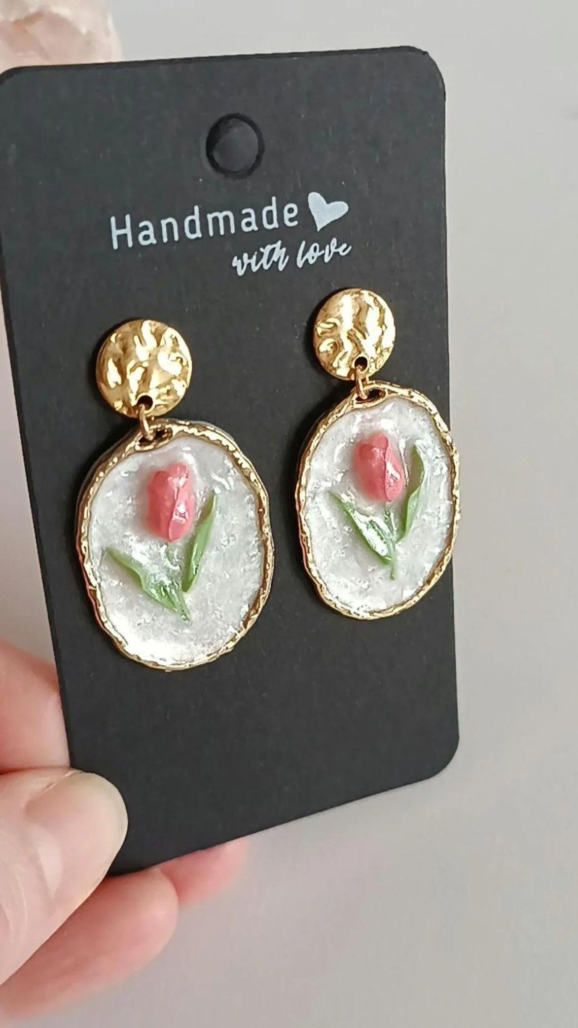 Close-up of handmade pink tulip dangle earrings hanging on a display card. The earrings are sculpted from polymer clay with green leaf details and a pearl accent