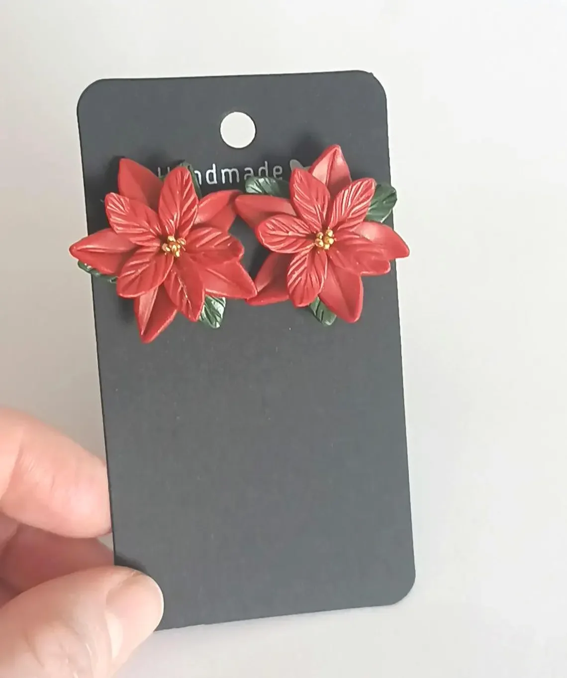 Festive red poinsettia stud earrings sculpted from polymer clay. The handmade floral studs feature green leaf details and stainless steel posts, displayed on a white background.