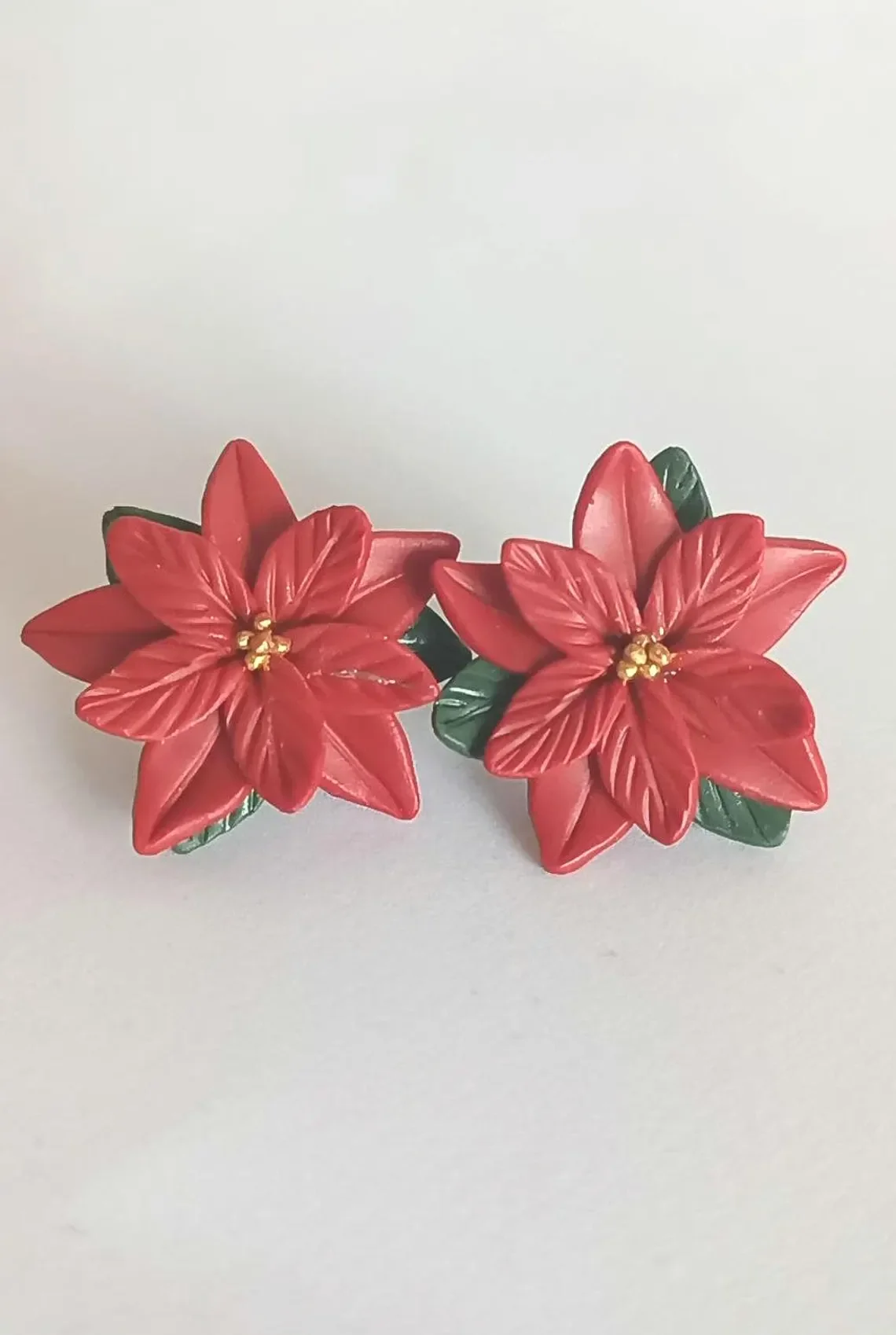 Festive red poinsettia stud earrings sculpted from polymer clay. The handmade floral studs feature green leaf details and stainless steel posts, displayed on a white background.