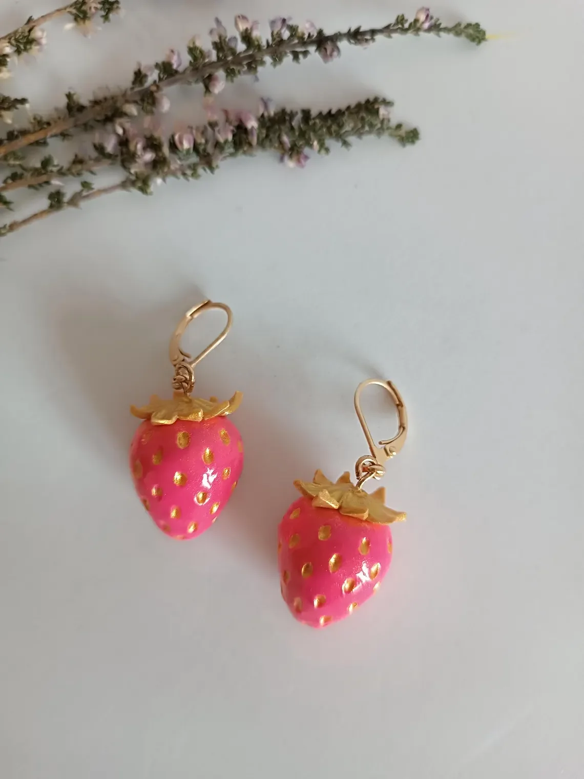 Pink polymer clay strawberry earrings with gold accents. Handmade fruit dangle earrings featuring gold-plated leaf caps and botanical details.