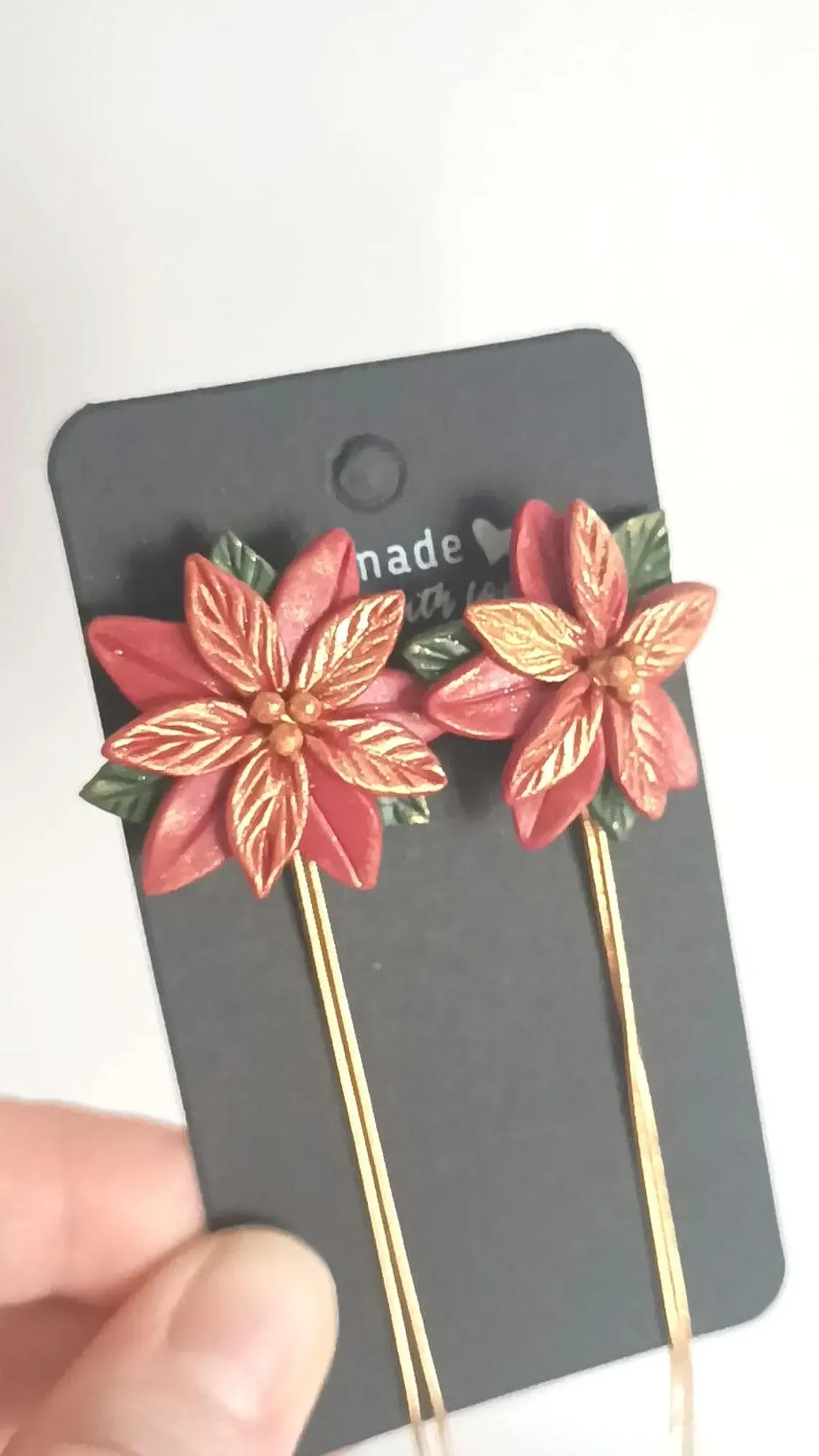 Festive red poinsettia stud earrings sculpted from polymer clay. The handmade floral studs feature green leaf details and stainless steel posts, displayed on a white background.