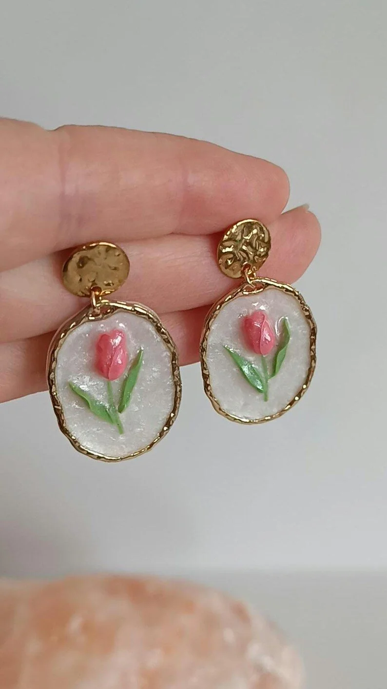Close-up of handmade pink tulip dangle earrings hanging on a display card. The earrings are sculpted from polymer clay with green leaf details and a pearl accent