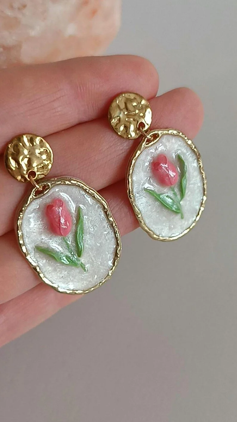 Close-up of handmade pink tulip dangle earrings hanging on a display card. The earrings are sculpted from polymer clay with green leaf details and a pearl accent
