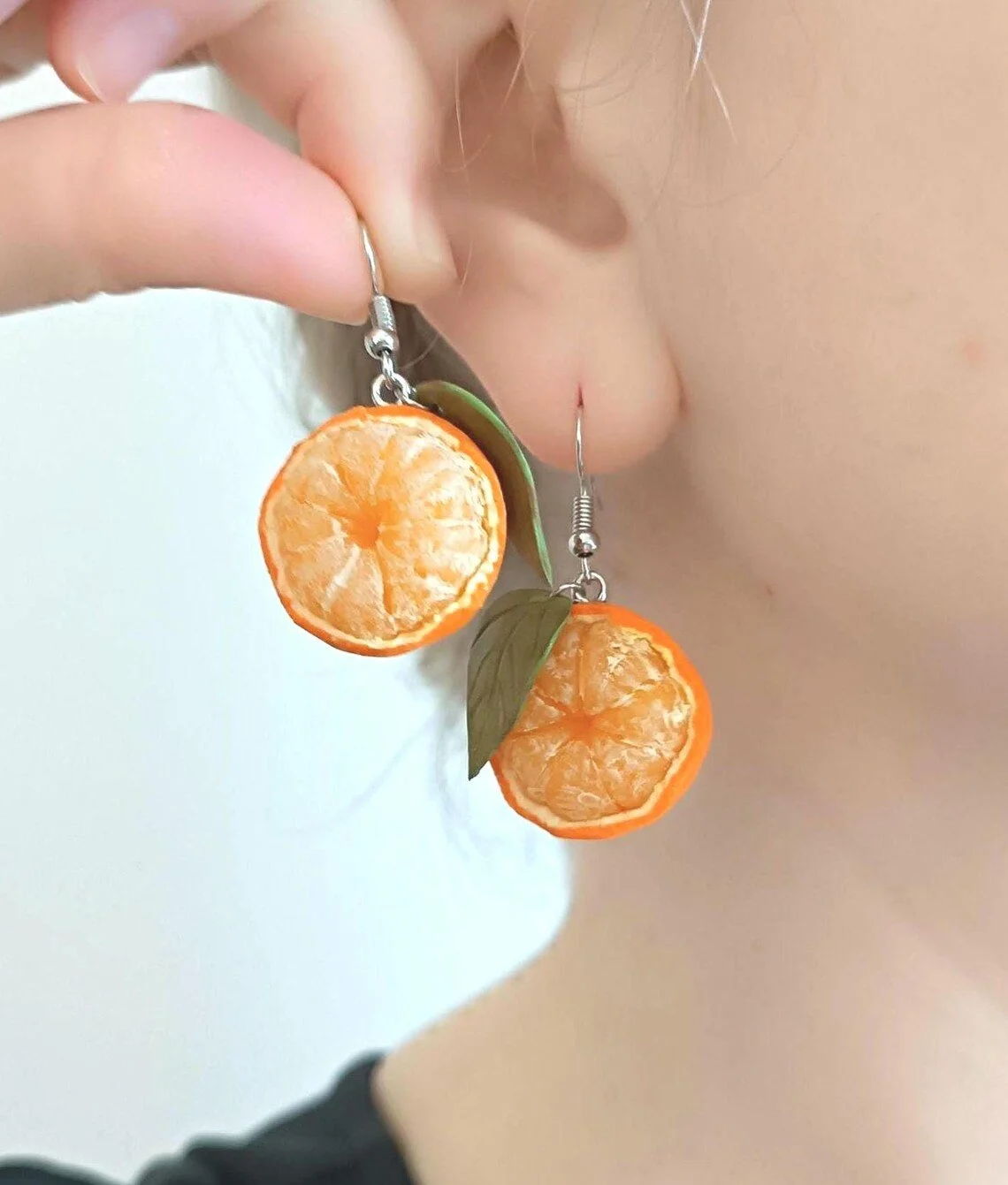 Earrings shaped like peeled oranges with green leaves, worn by a person near their ear.