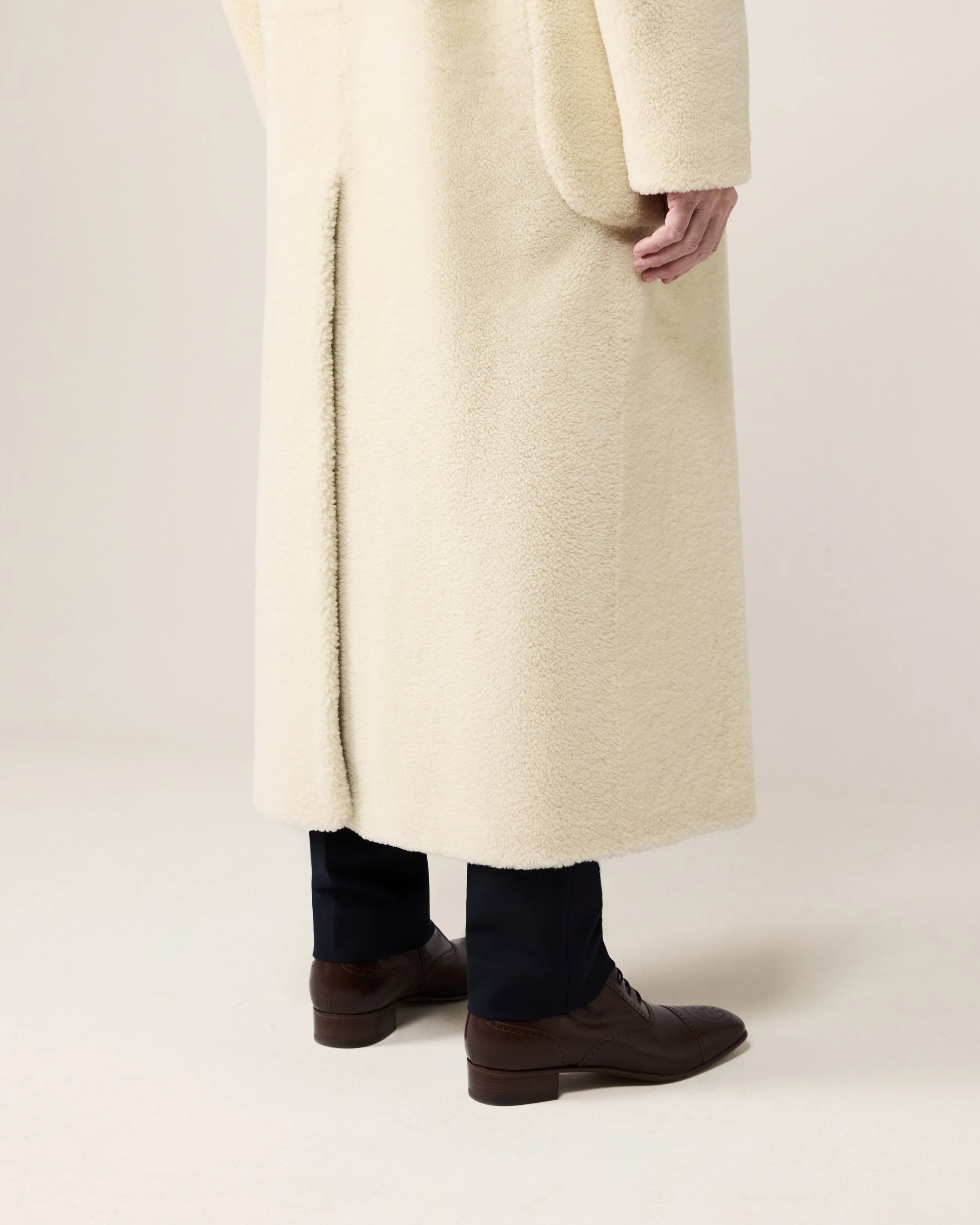 Nahaufnahme von den unteren Körperteilen einer Person, die hellbeige, lange Wollmantel trägt, braune Lederschuhe und dunkle Hosen. Der Hintergrund ist weiß.