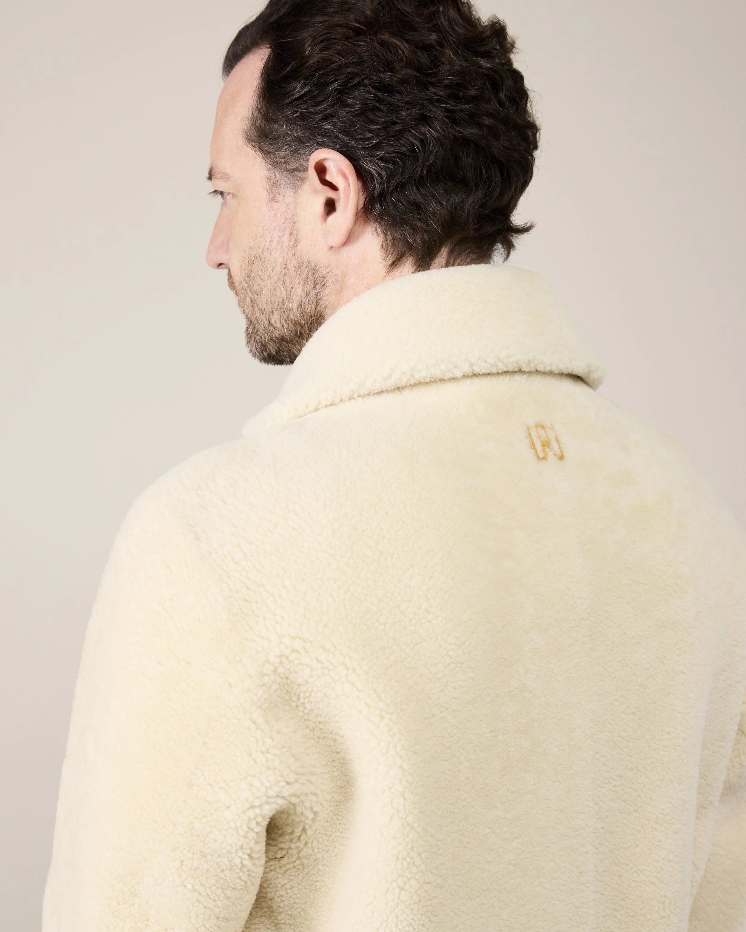 Ein Mann trägt einen beigen, ,Sherpa-ähnlichen' Pullover mit hochstehendem Kragen, nach hinten gedreht, zeigt den Rücken. Das Material ist wollig und kuschelig.