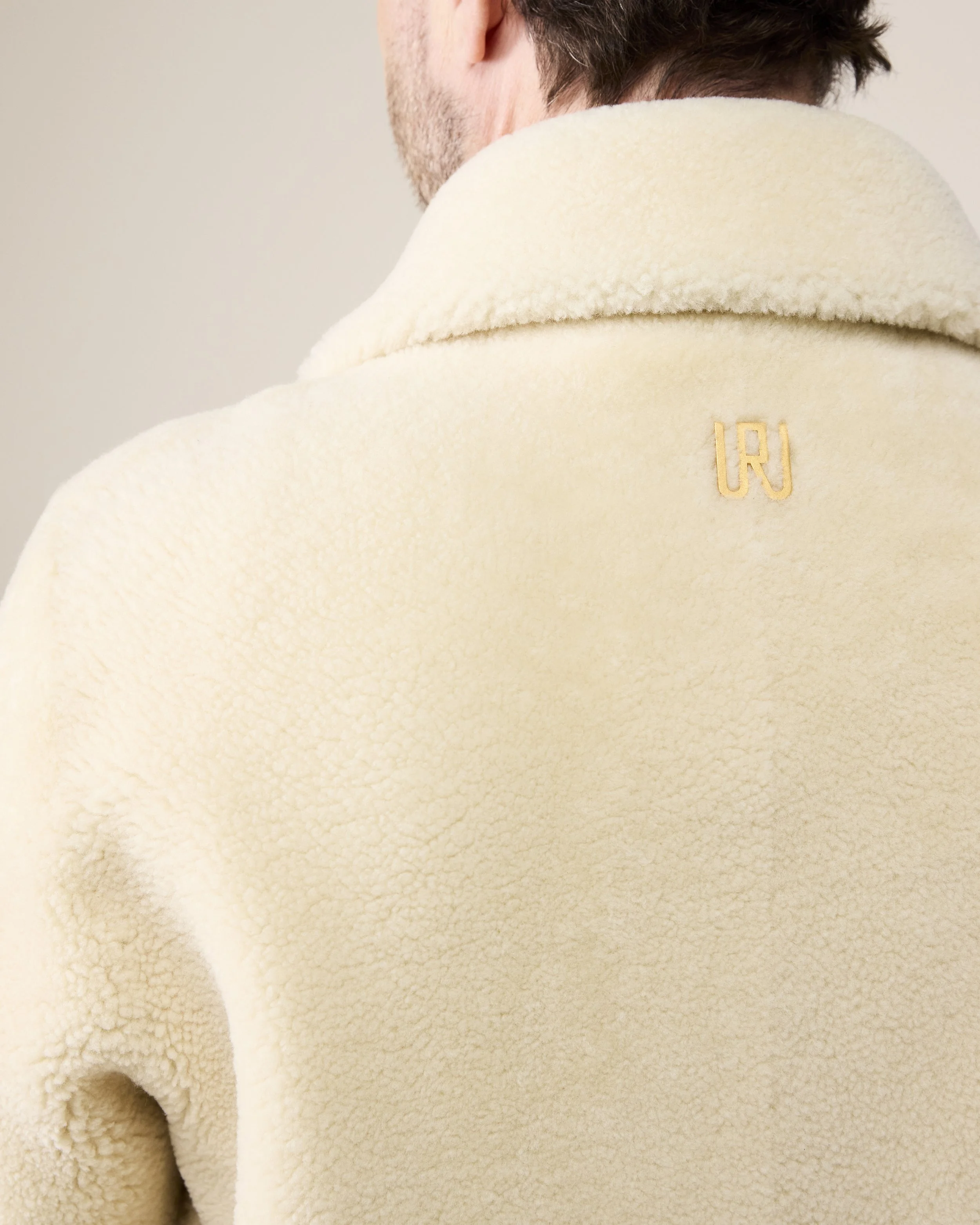 Nahaufnahme eines Mannes, der einen cremefarbenen, flauschigen Pullover trägt, mit einem kleinen goldenen Logo auf dem Rücken