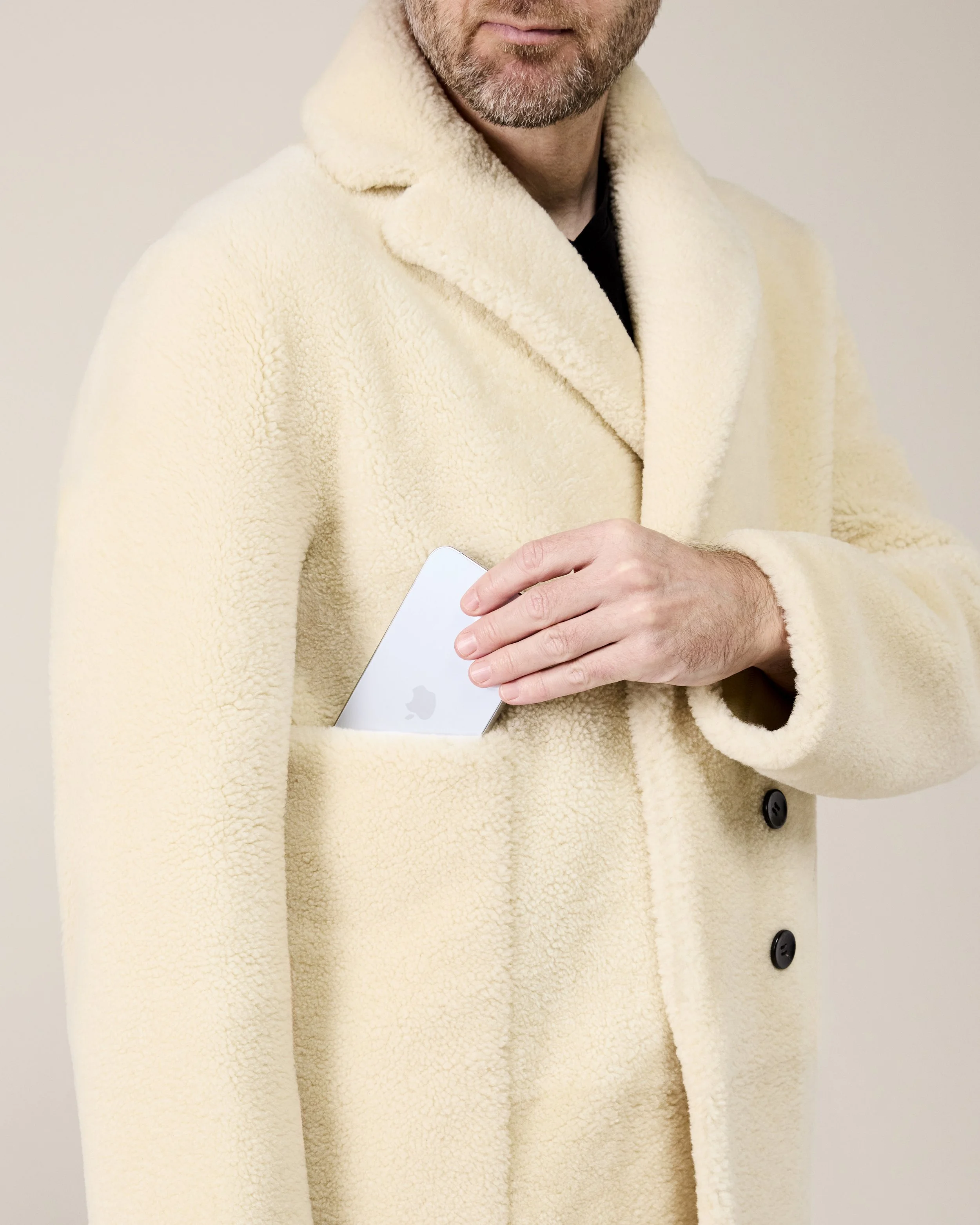 Ein Mann in einem cremefarbenen Pelzmantel steckt ein iPhone in die Manteltasche.