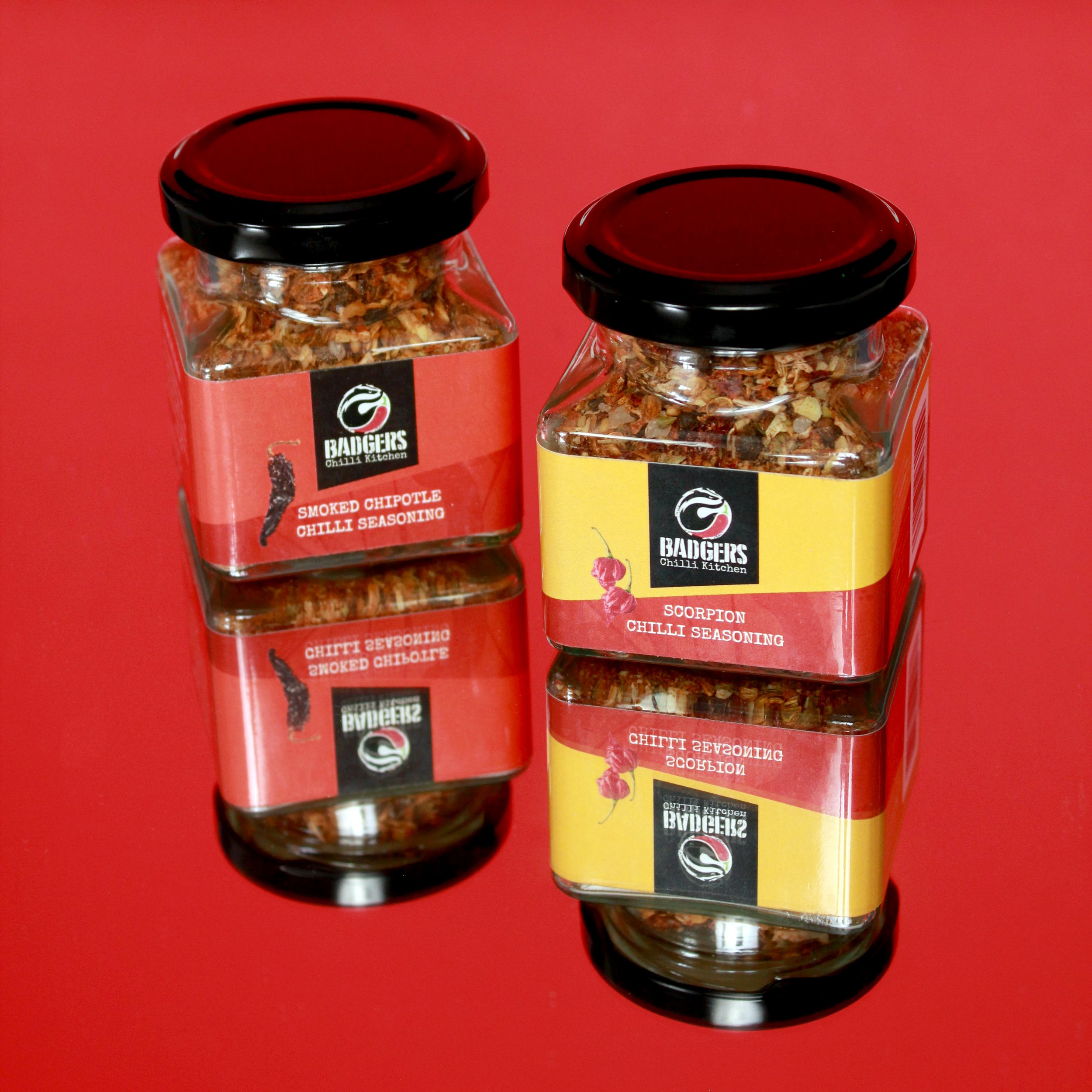 Two jars of chili seasoning from Badgers Chilli Kitchen, one labeled smoked chipotle and the other scorpion, on a red reflective surface.