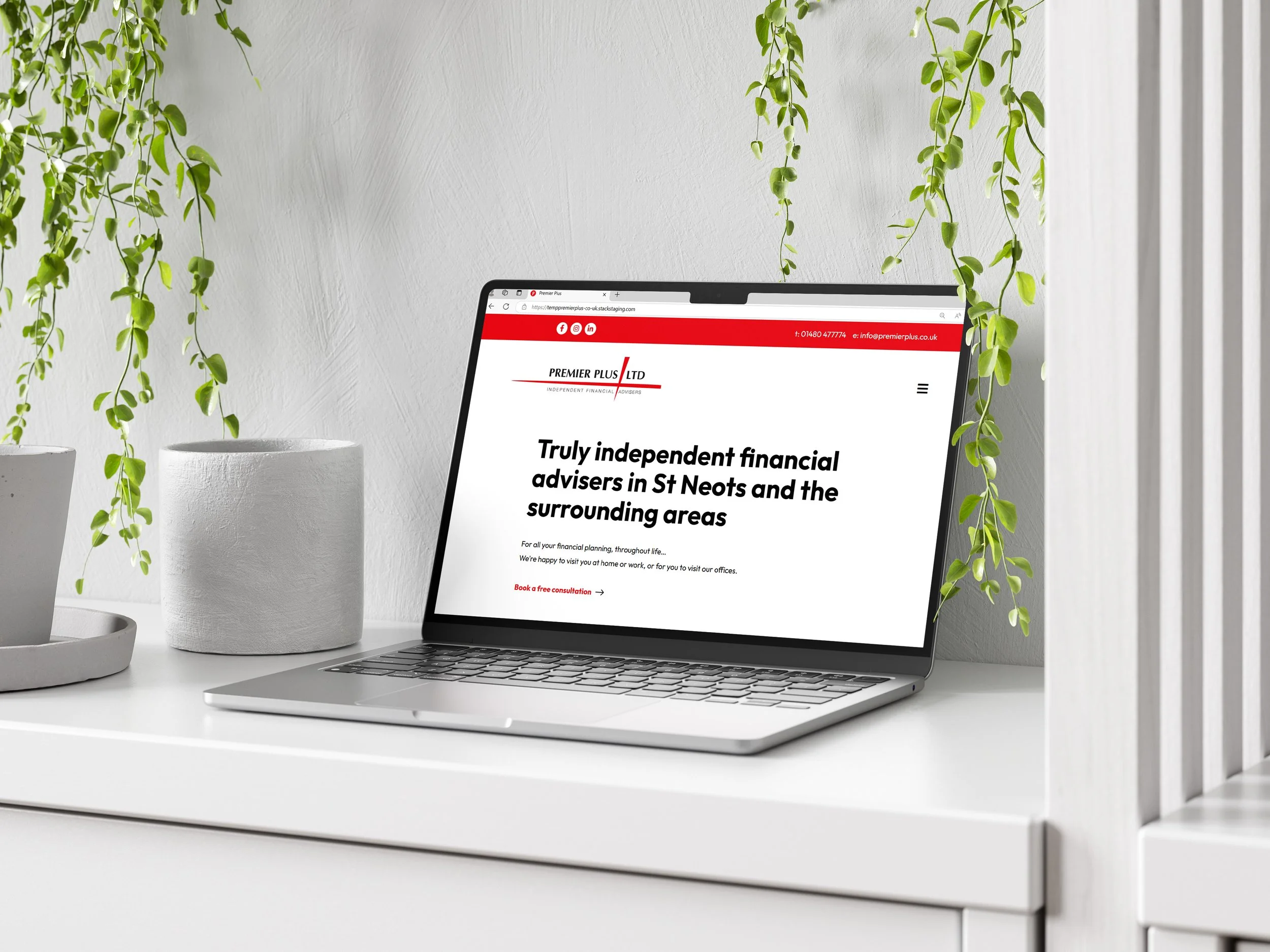 Open laptop on a white desk displaying a financial advisory website, with white pots of plants and hanging green plants in the background.