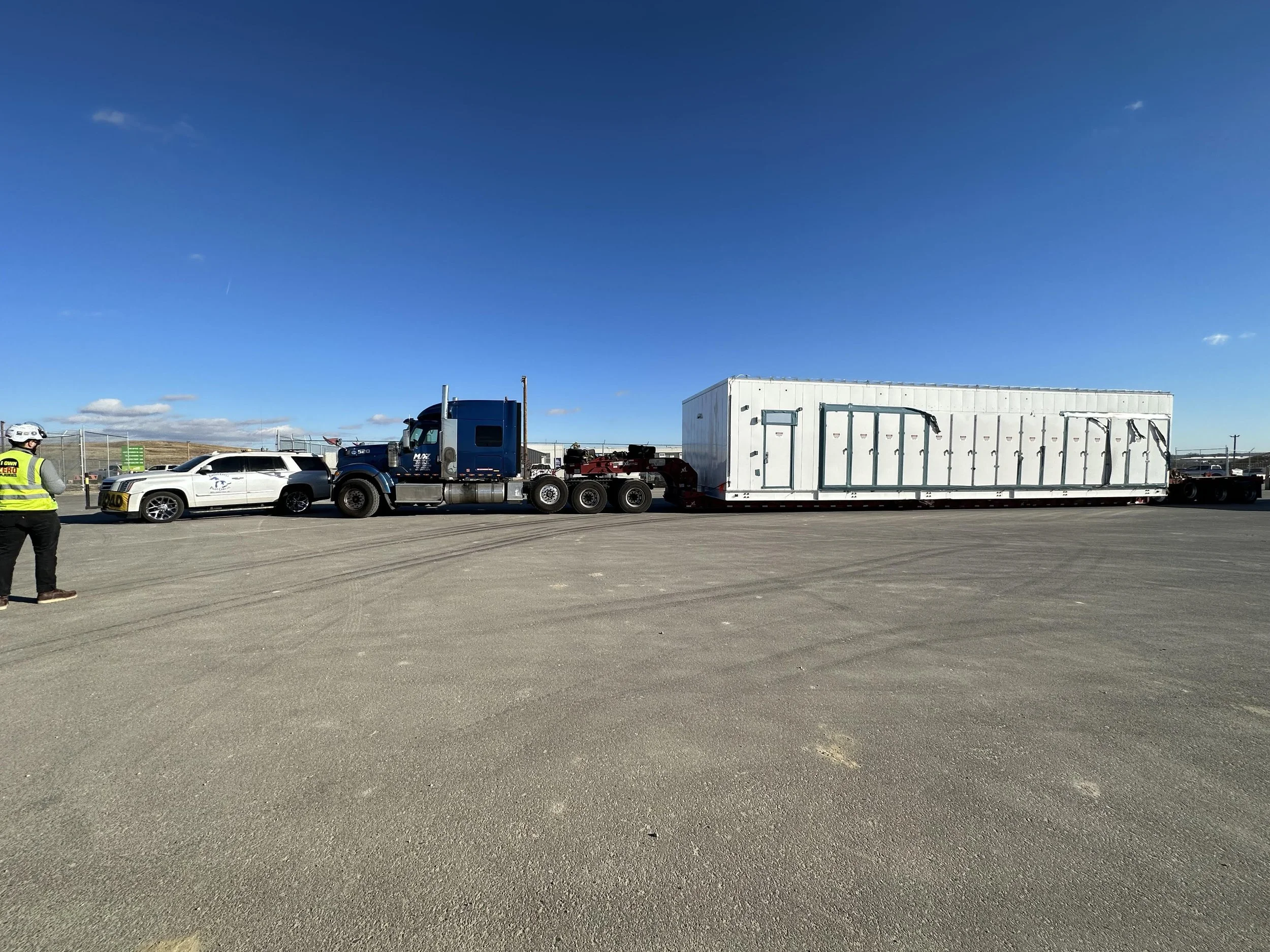 A large semi-truck transporting a white container-like structure on a flatbed trailer, with a white car and a person in a safety vest and helmet nearby.